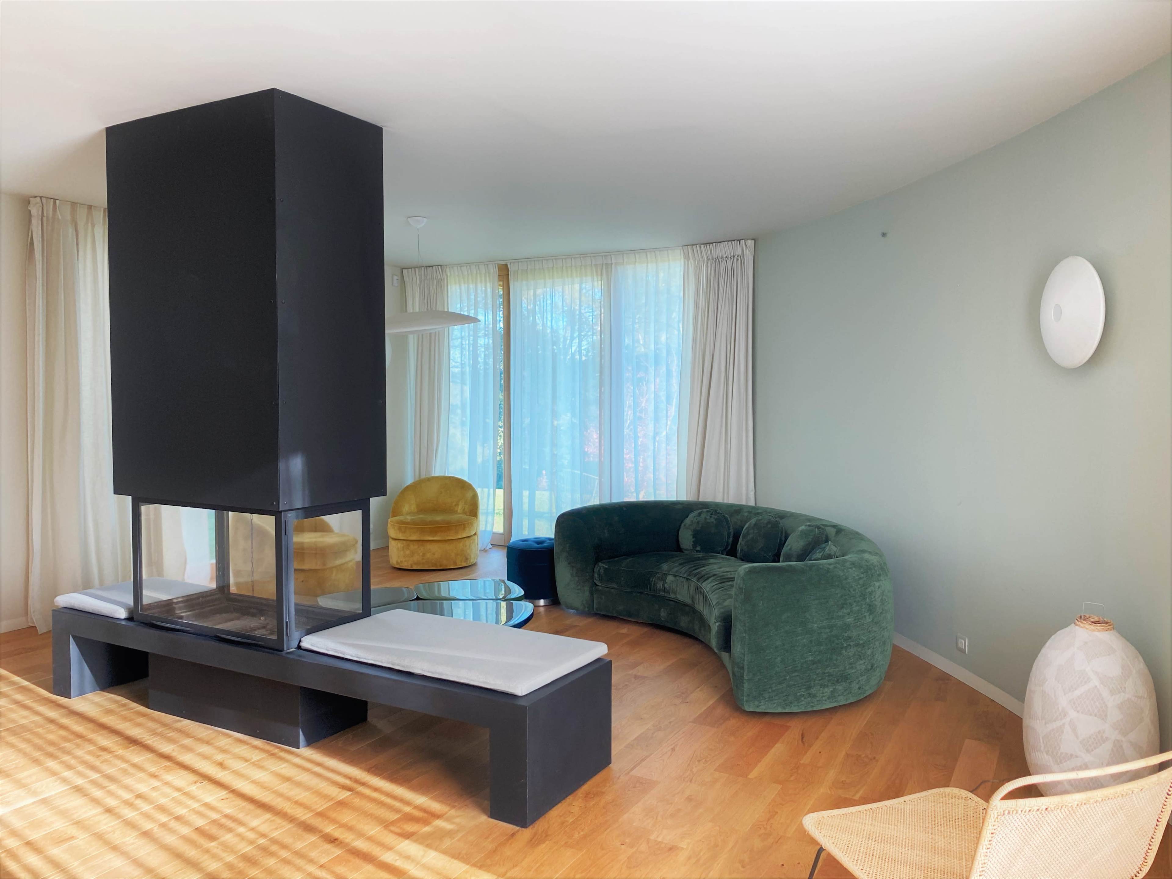 A modern living room featuring a central black fireplace, a curved green sofa, a yellow armchair, and large windows letting in natural light.
