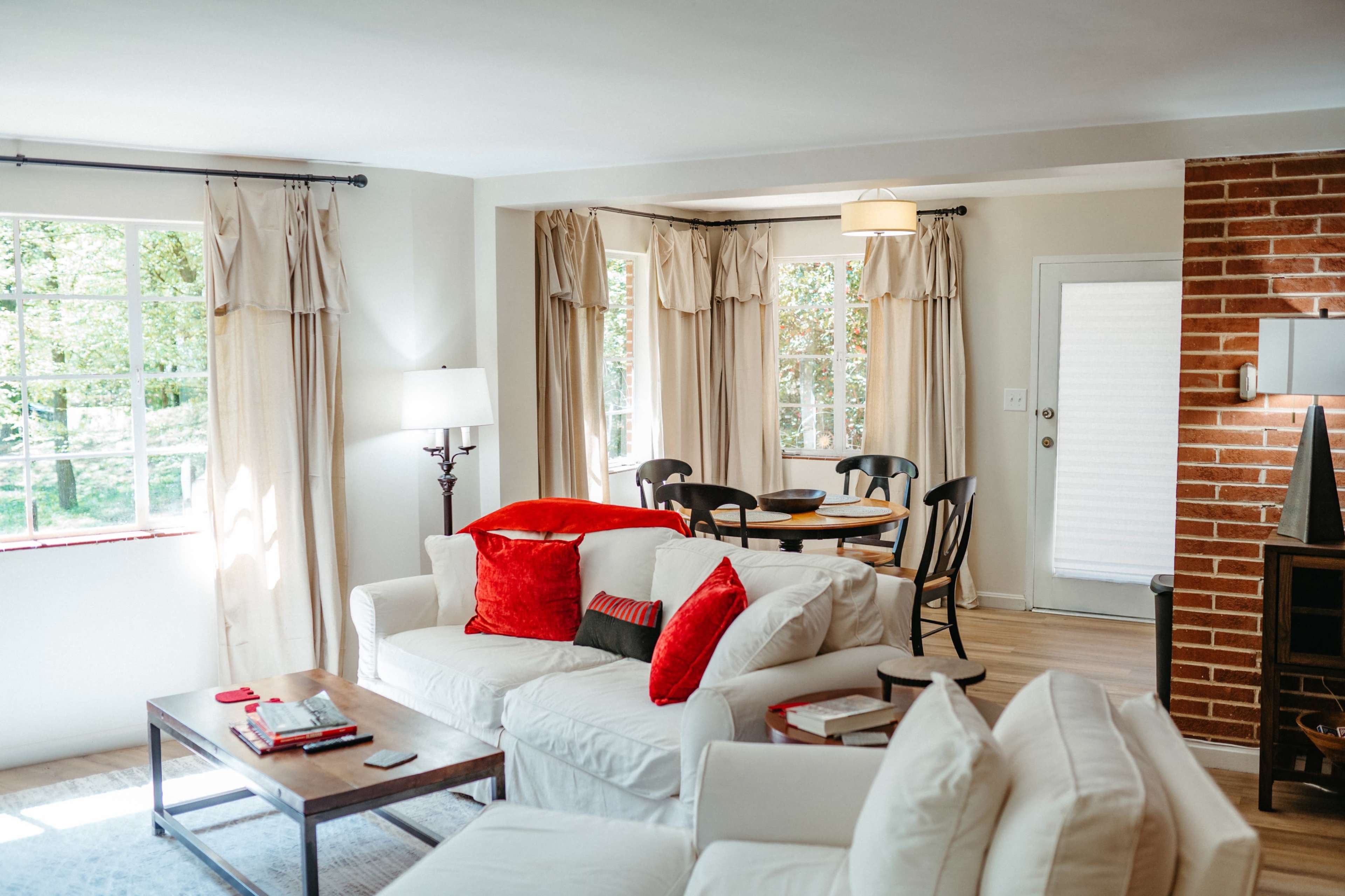 The image shows a bright living room with white sofas, a wooden coffee table, and a dining area featuring a round table and chairs, complemented by large windows with light curtains.