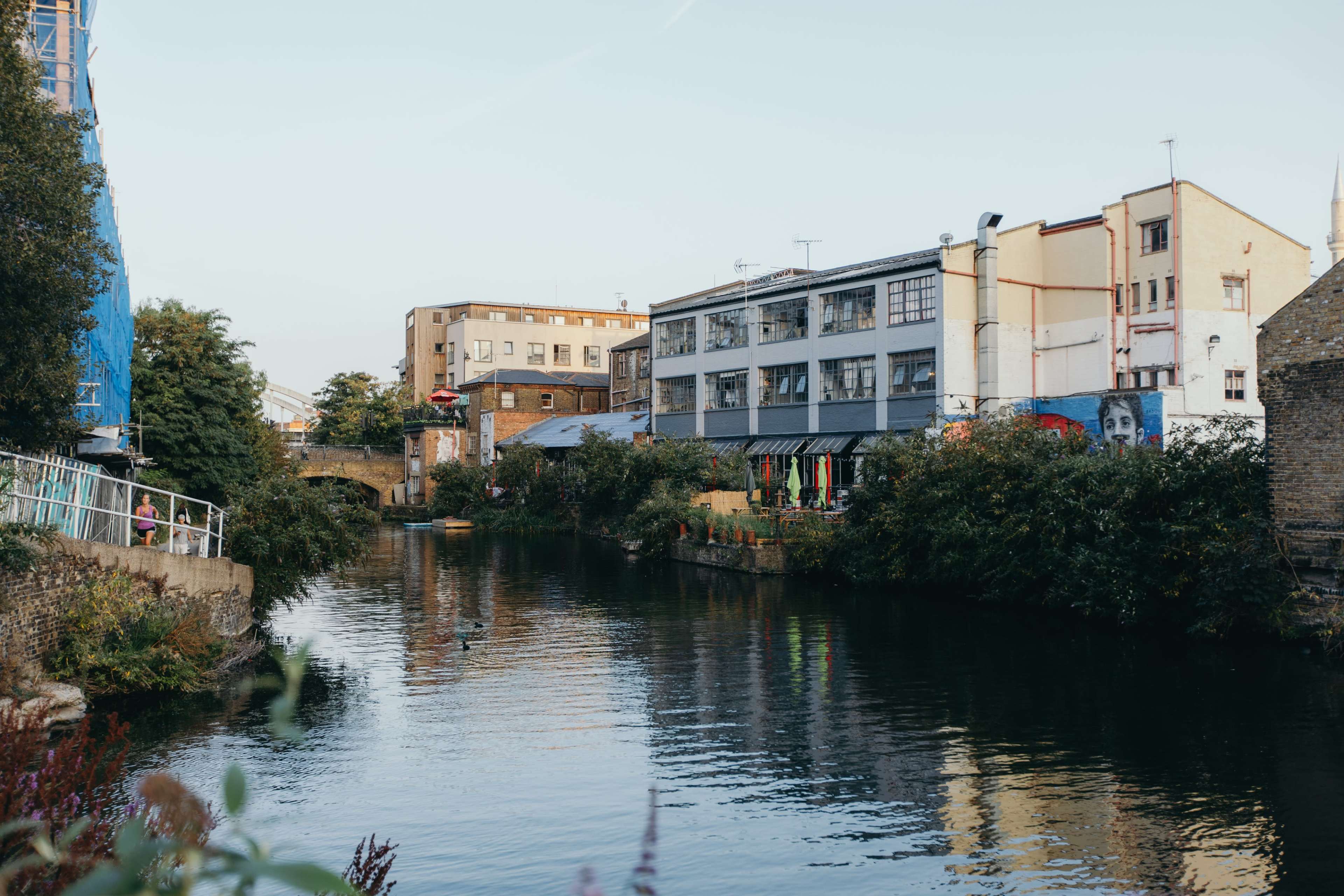 A canal lined with buildings and greenery, reflecting a clear sky above.