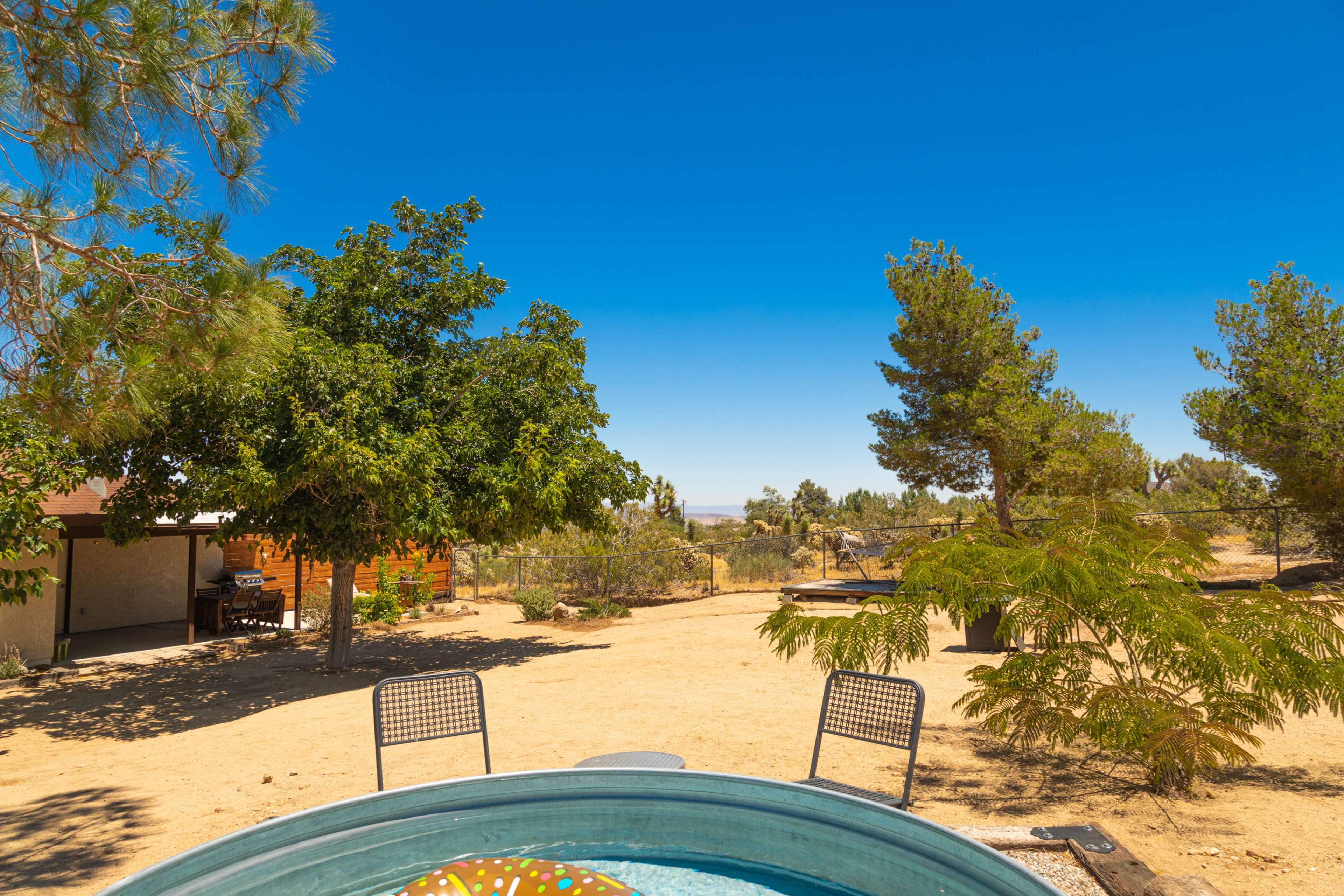 A hot tub sits surrounded by dry landscaping, with trees and a distant building visible under a clear blue sky.