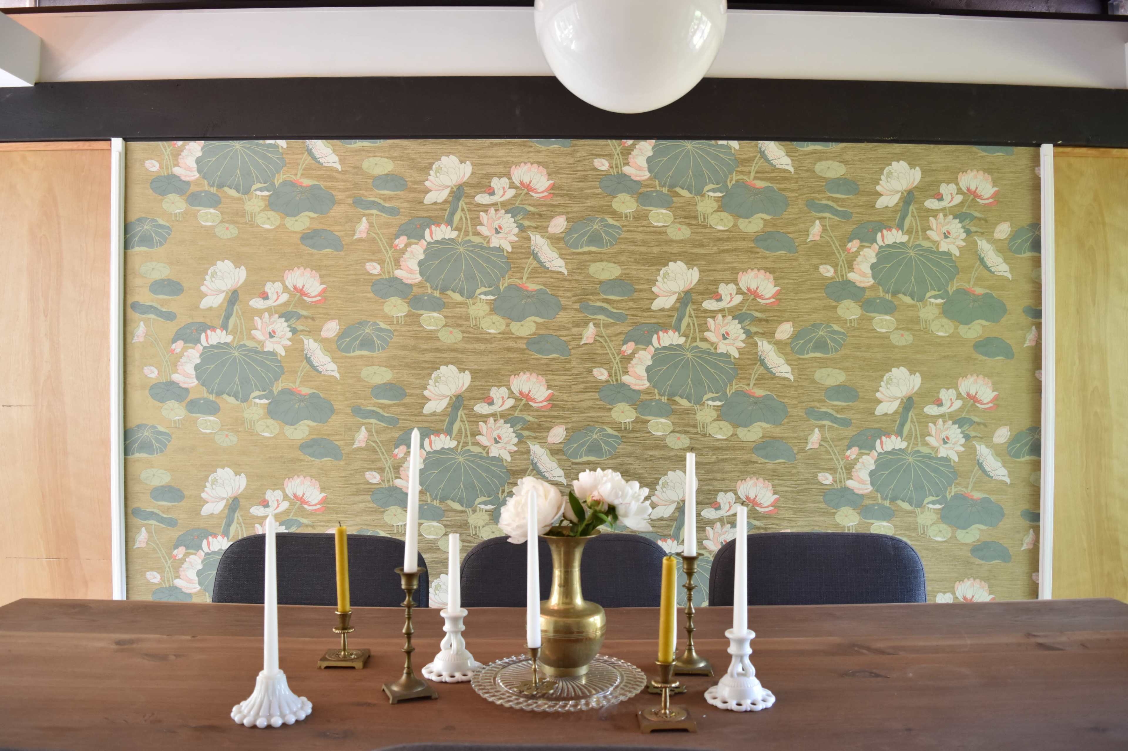 A wooden dining table is set with various candle holders and a vase of flowers against a decorative lotus-patterned wall.