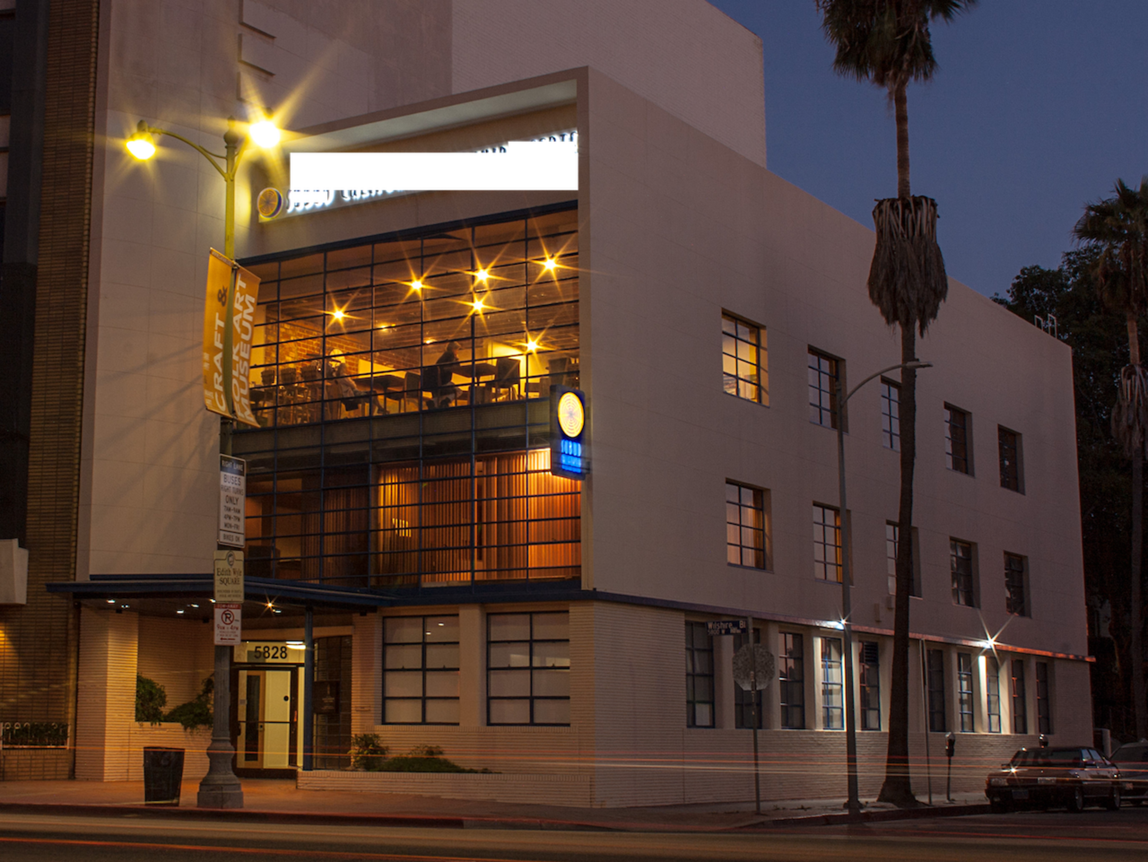 The image shows a modern building with large windows and illuminated interior spaces, located on a street lined with palm trees at dusk.
