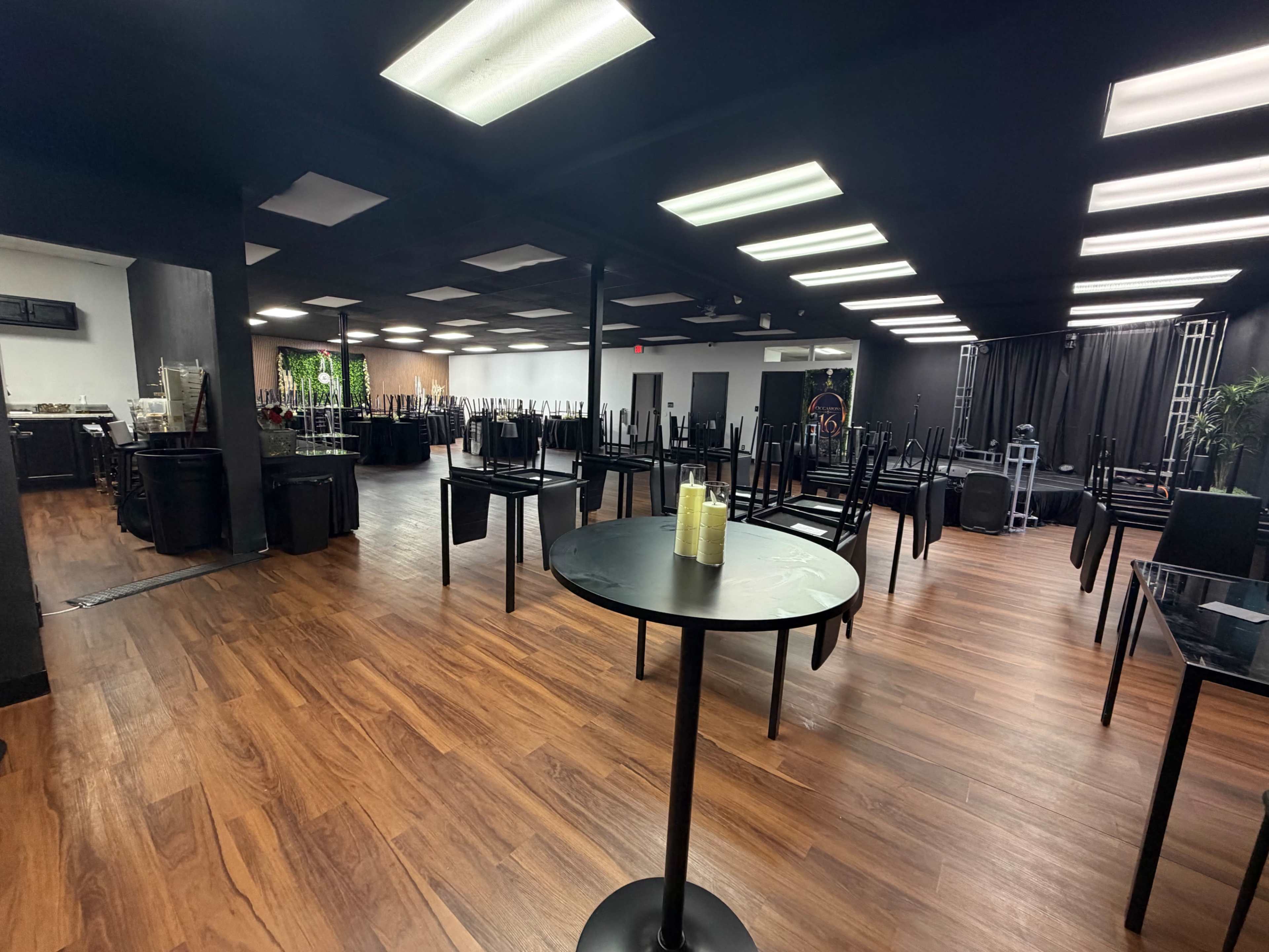 The image shows an empty event space with tables stacked upside down, chairs placed on top of the tables, and a dark floor with overhead lighting.