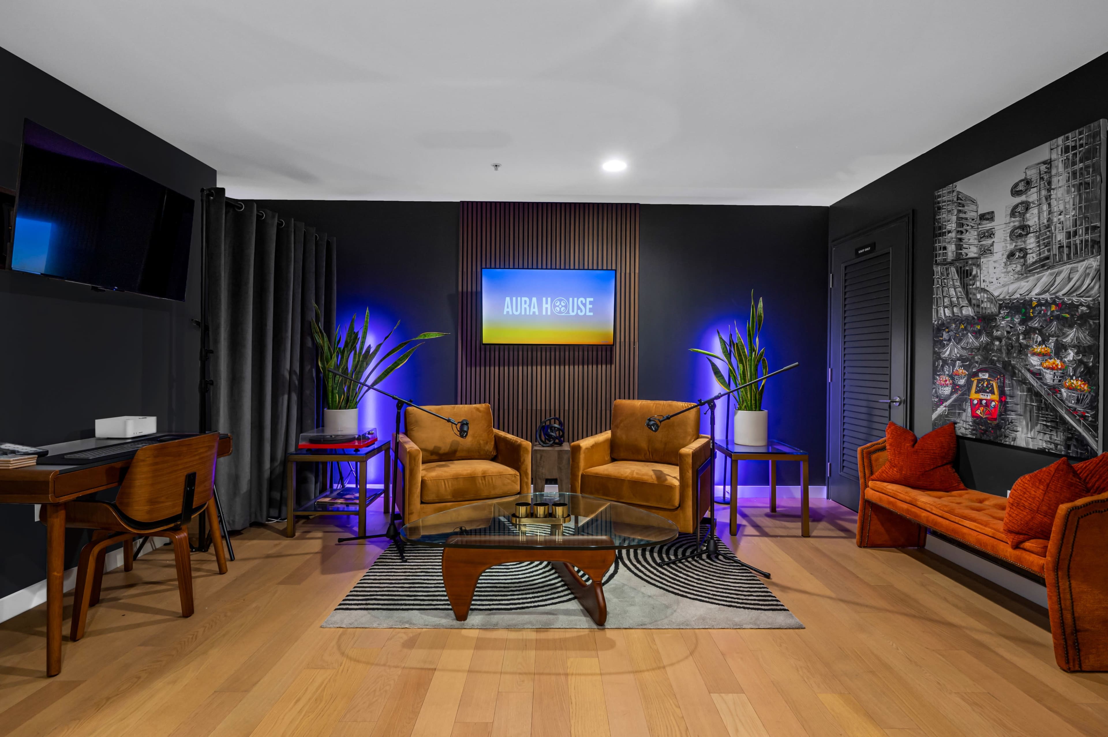 A modern lounge area featuring two orange chairs, a red sofa, a glass coffee table, and a wall-mounted screen with ambient lighting.