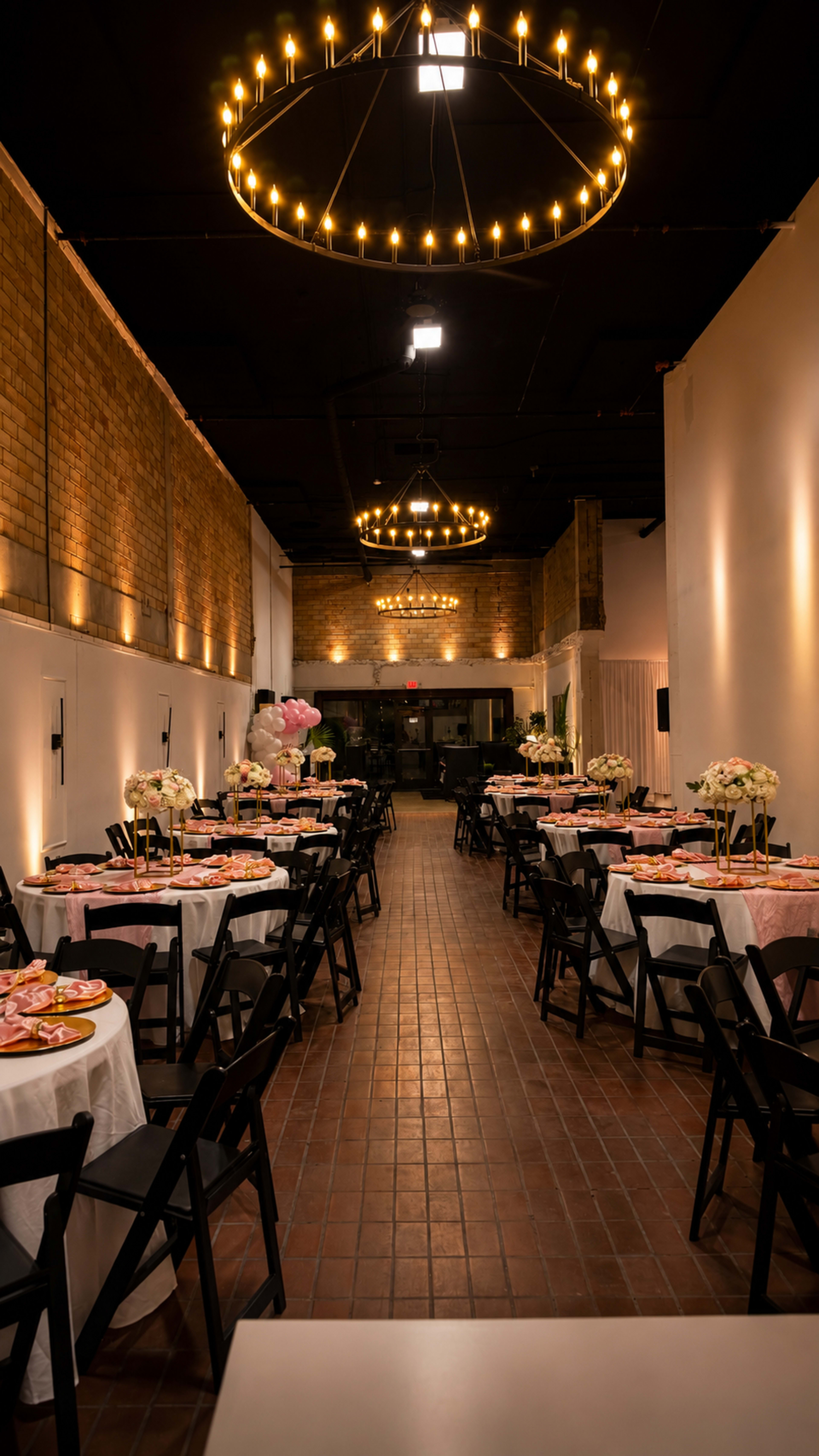 The image shows a spacious event room with elegantly set round tables, each adorned with pink plates and floral centerpieces, under decorative chandelier lighting.