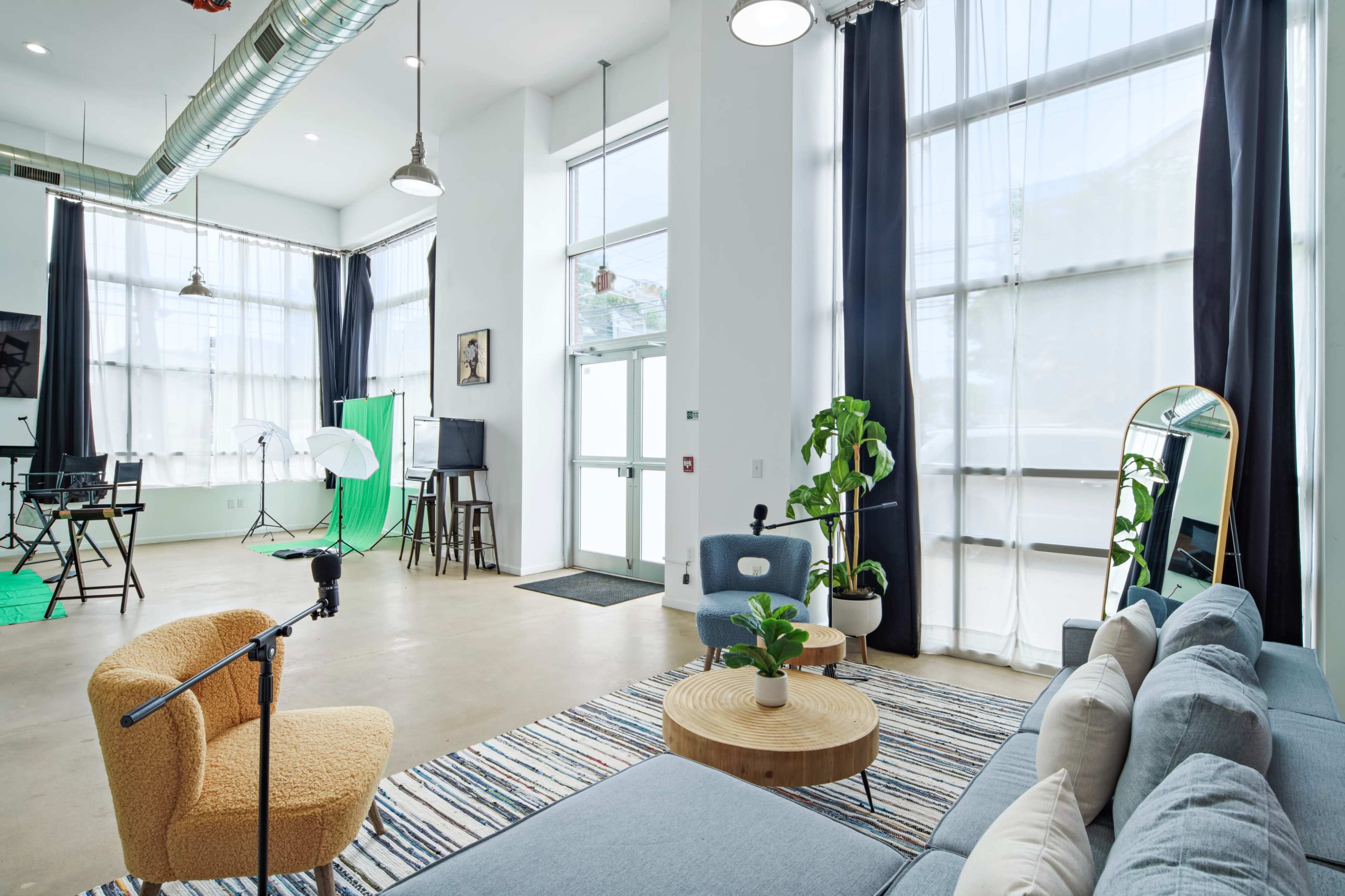 A bright, modern studio space with large windows, a combination of seating areas, and a green backdrop setup for photo or video production.