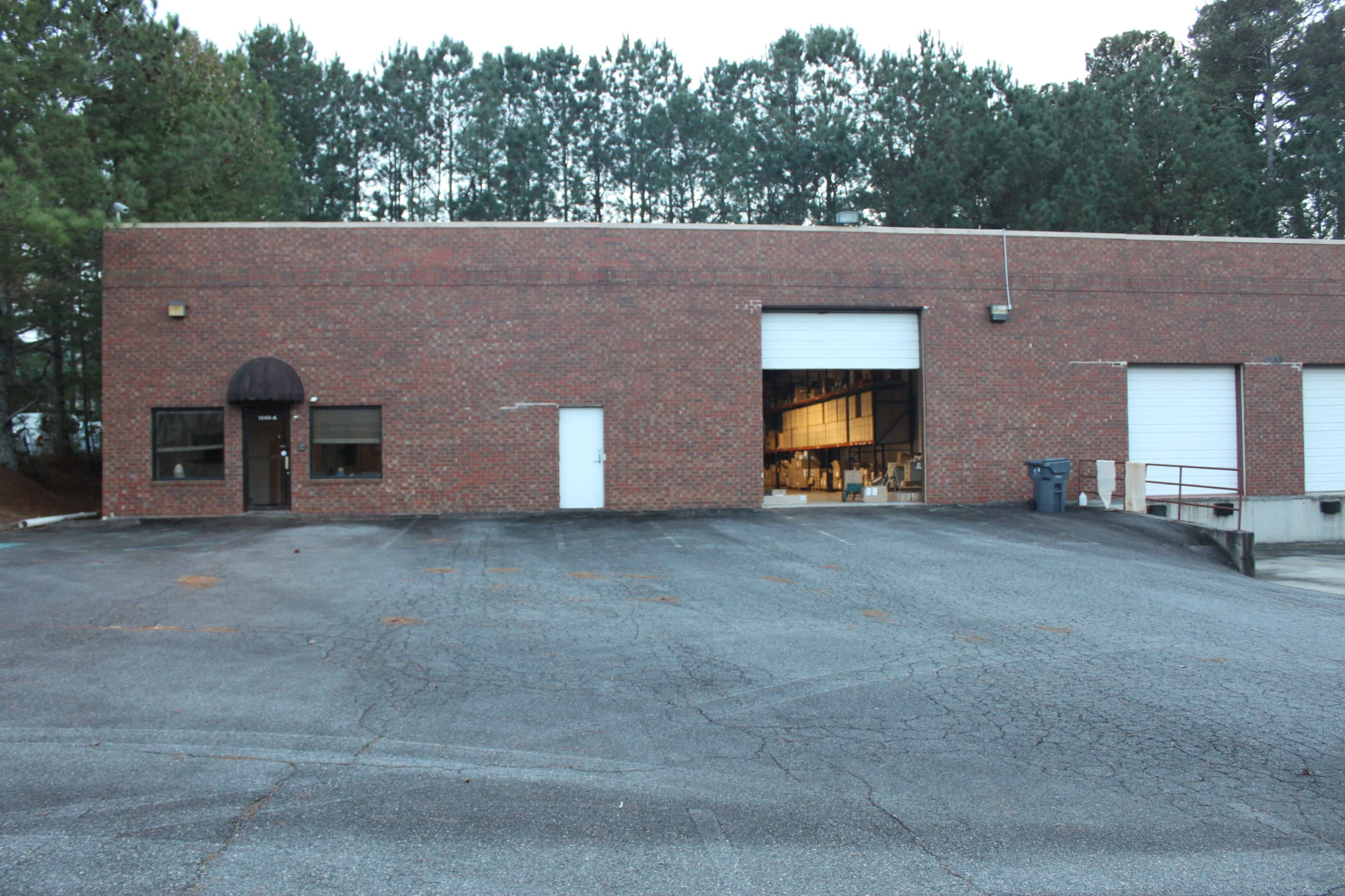 The image shows a brick warehouse building with large loading doors and a paved area in front, surrounded by trees in the background.