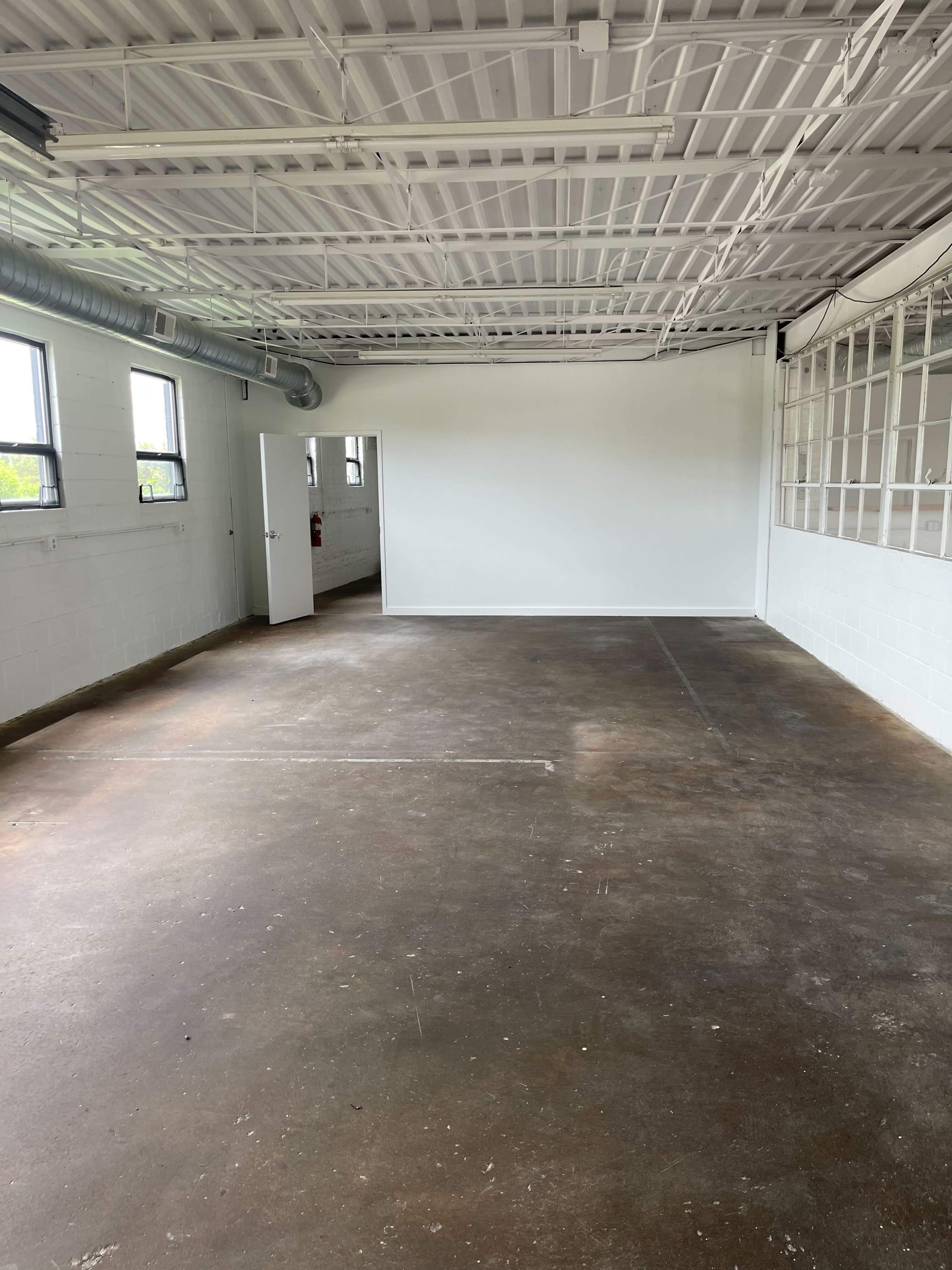 An empty industrial-style room with concrete flooring, white walls, and large windows.