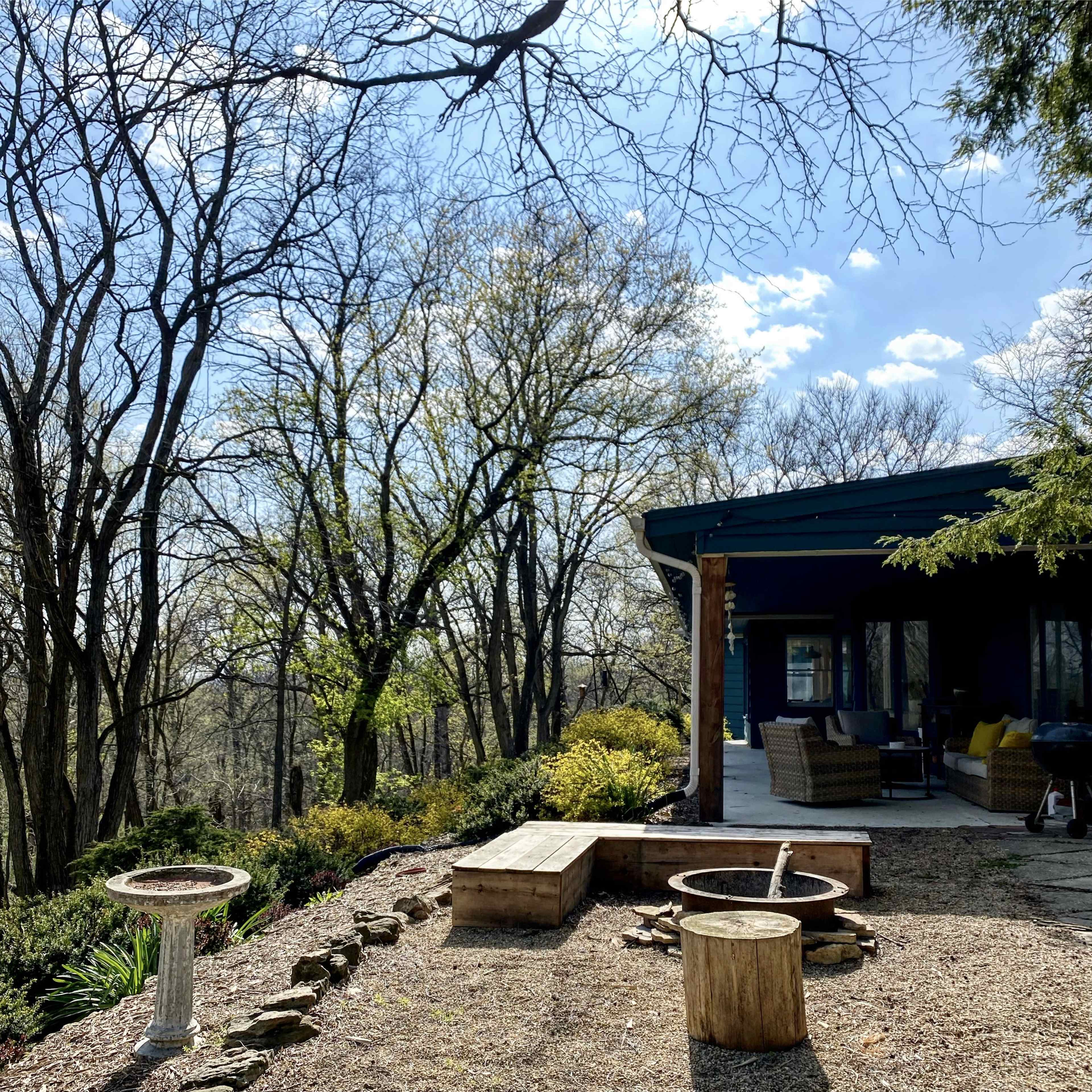 A patio area with wooden seating, a fire pit, and a birdbath, surrounded by trees and greenery under a clear blue sky.