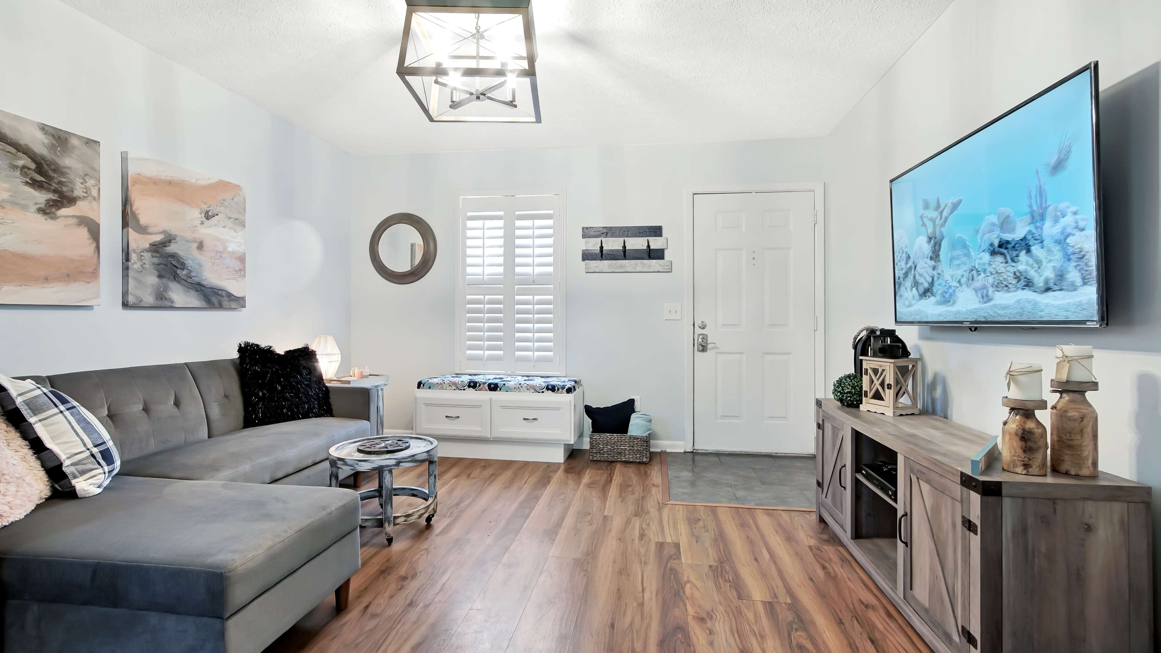 A living room with a gray sectional sofa, a wooden coffee table, a television mounted on the wall, and artwork hanging on the light-colored walls.