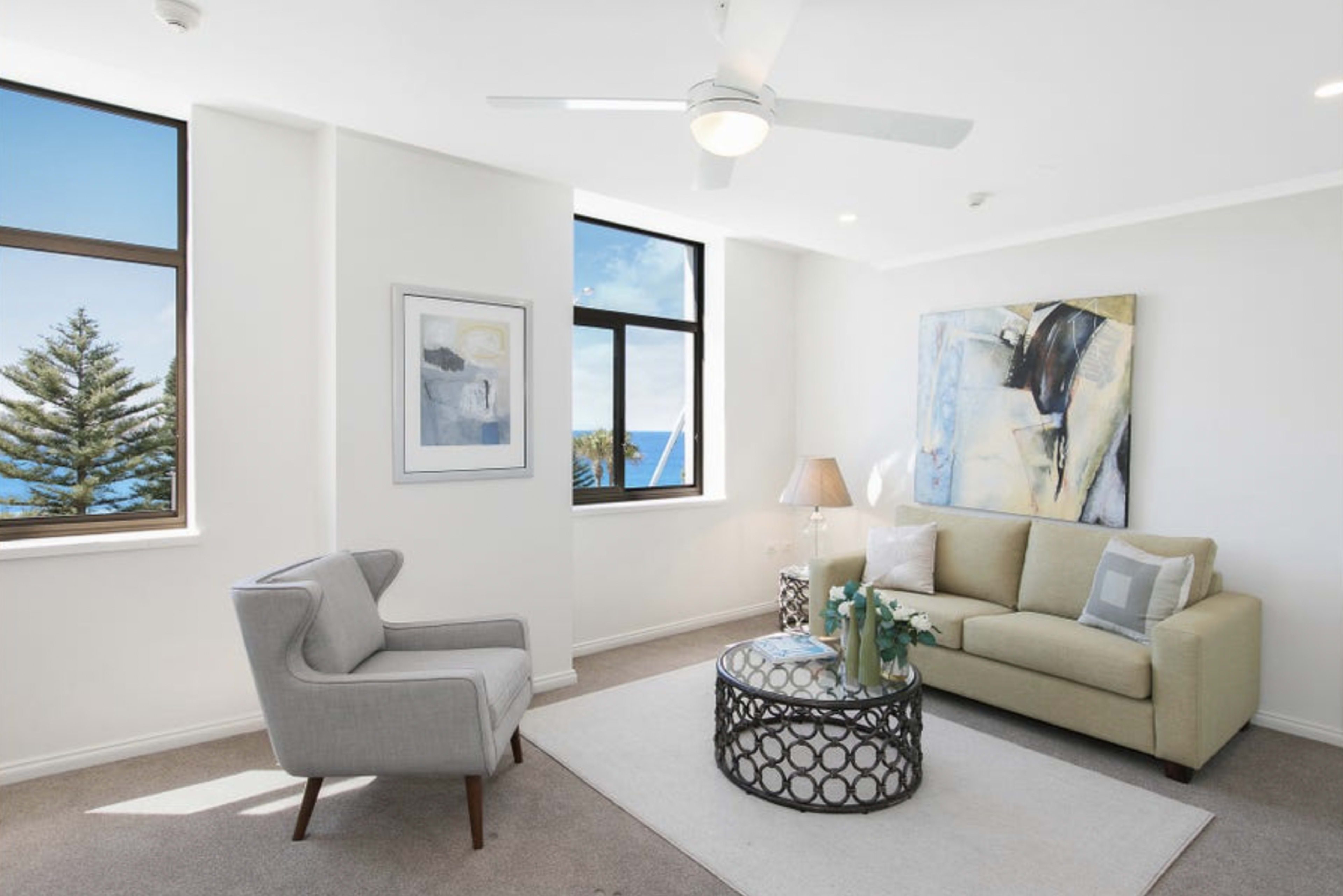 A bright living room features a light-colored sofa, a patterned coffee table, and large windows with a view of trees and the ocean.
