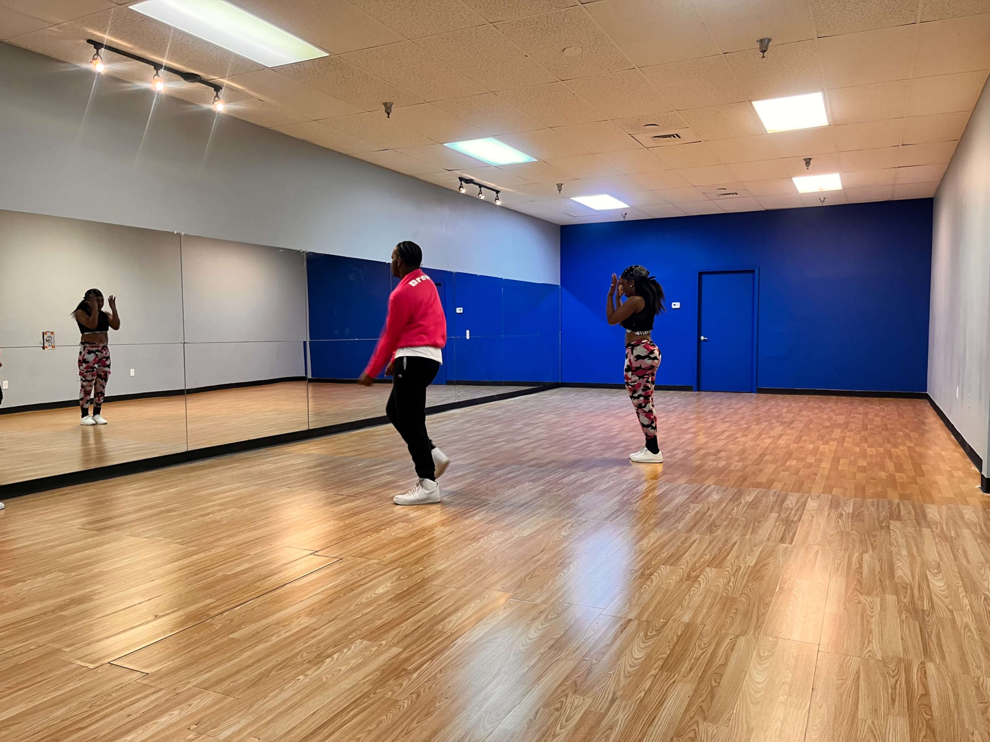 Two dancers practice in a mirrored dance studio with a blue accent wall and wooden flooring.
