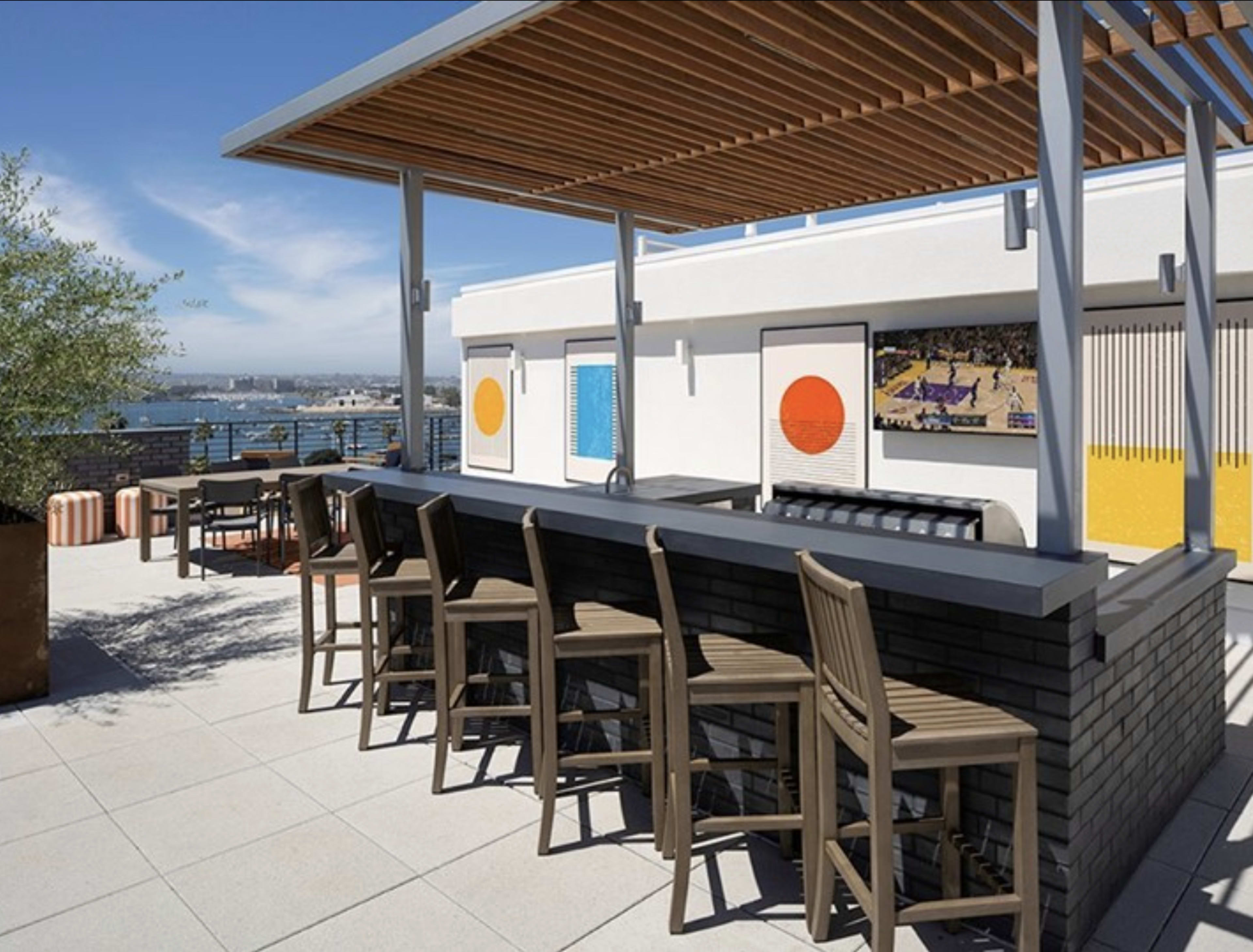 The image shows a rooftop bar with wooden seating and a view of a city skyline across the water.