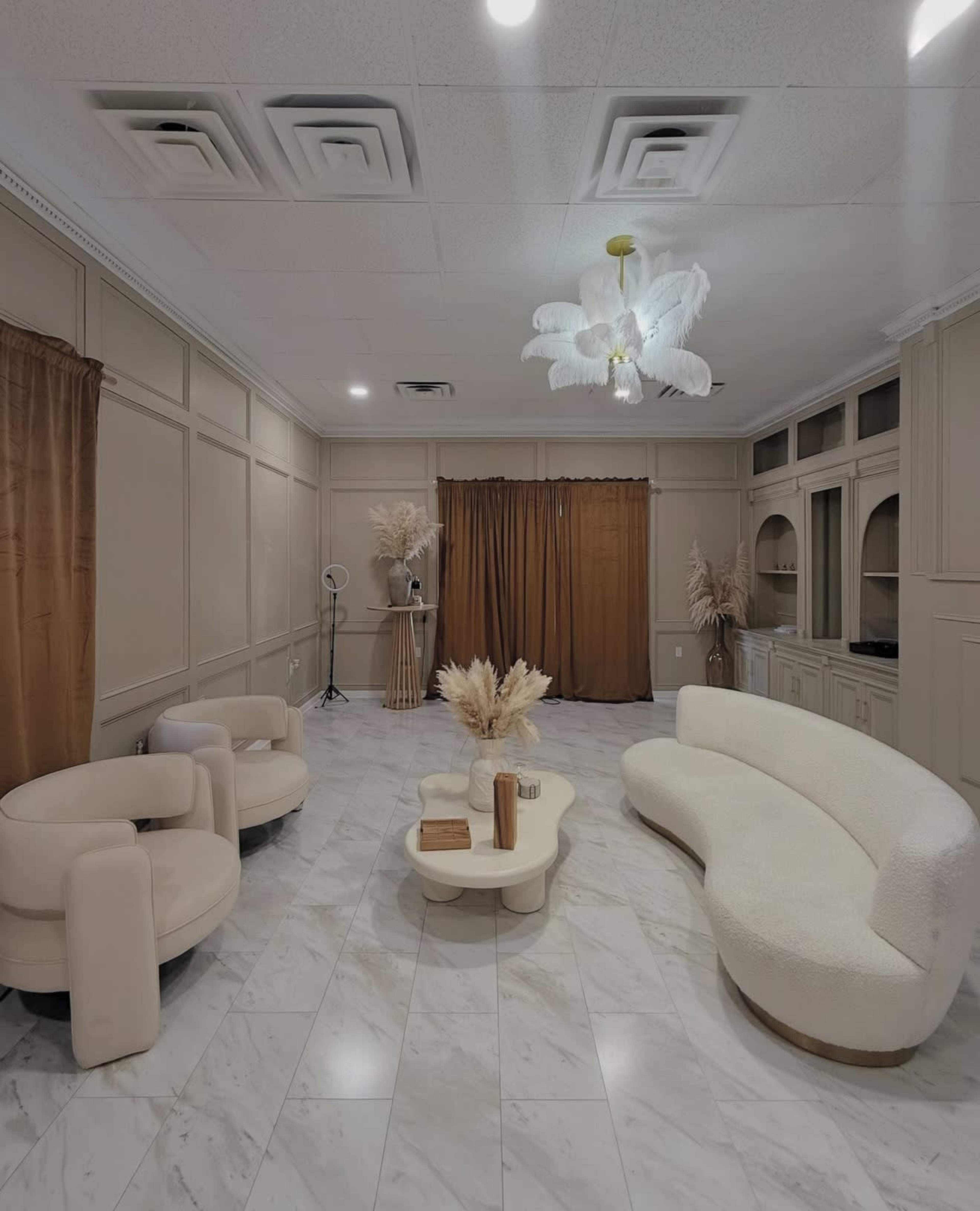 The image shows a modern lounge area with a curved white sofa, two circular chairs, a light-colored coffee table, and large brown curtains framing the space, all set against a marble floor.
