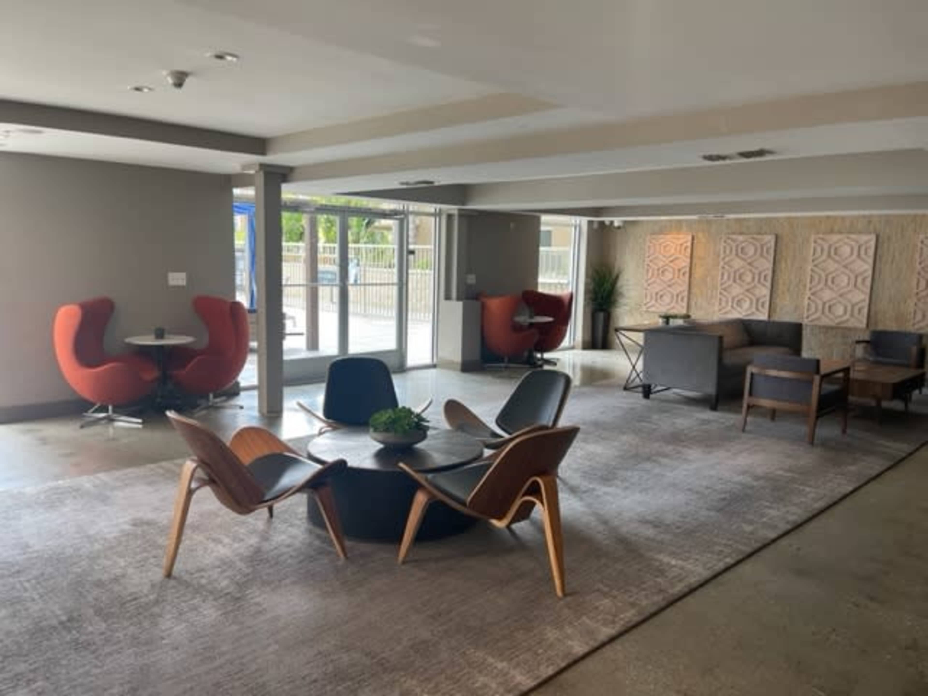 The image shows a modern lounge area featuring various seating arrangements, including orange chairs and a circular table, under natural lighting.