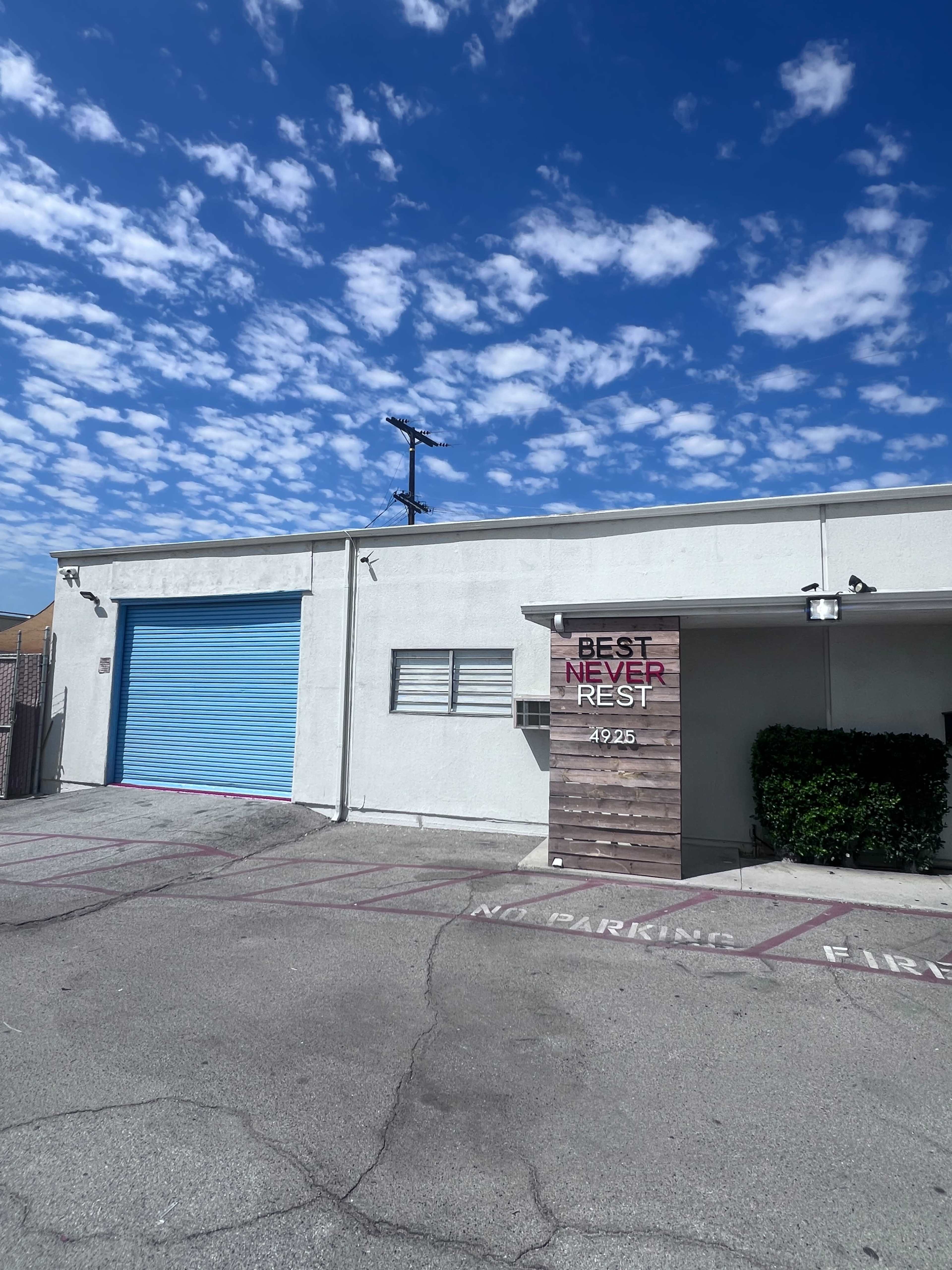 A white building with a blue garage door features a wooden sign that reads "BEST NEVER REST" on the exterior wall.