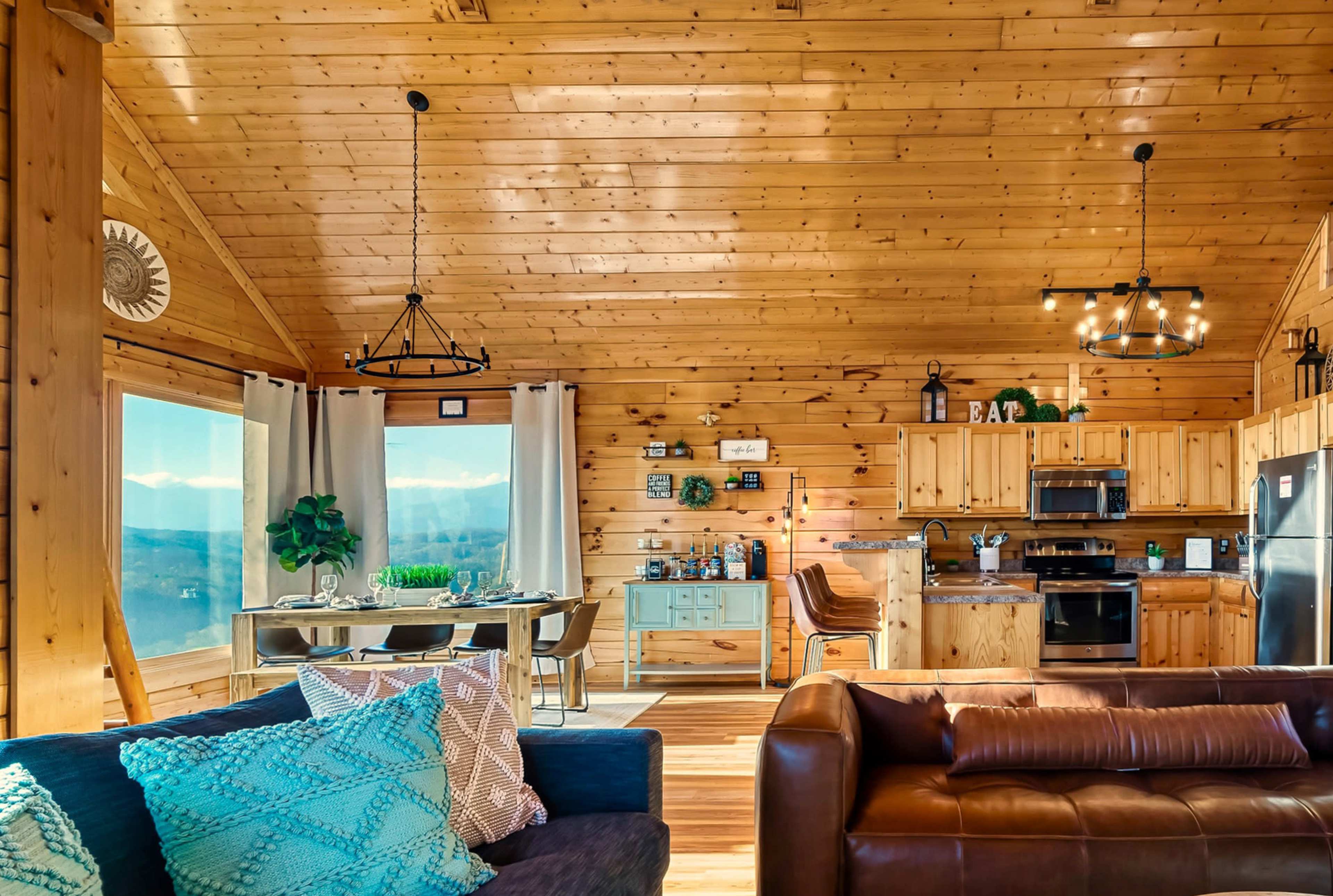 The image shows a wooden cabin interior with a kitchen and dining area, featuring large windows that reveal mountain views.