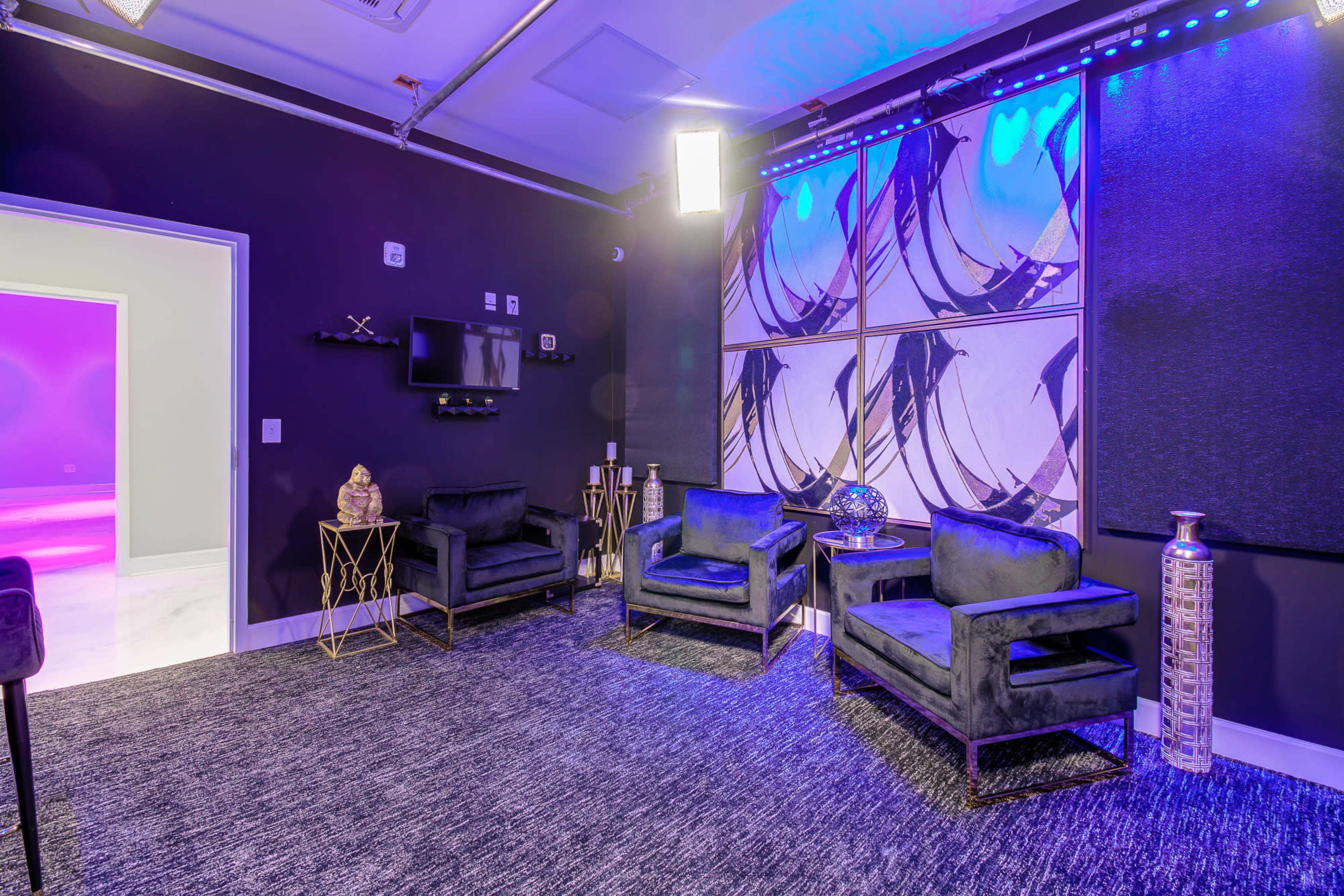 The image shows a modern lounge area with two dark velvet chairs, a small table, and abstract wall art, all illuminated by purple lighting.