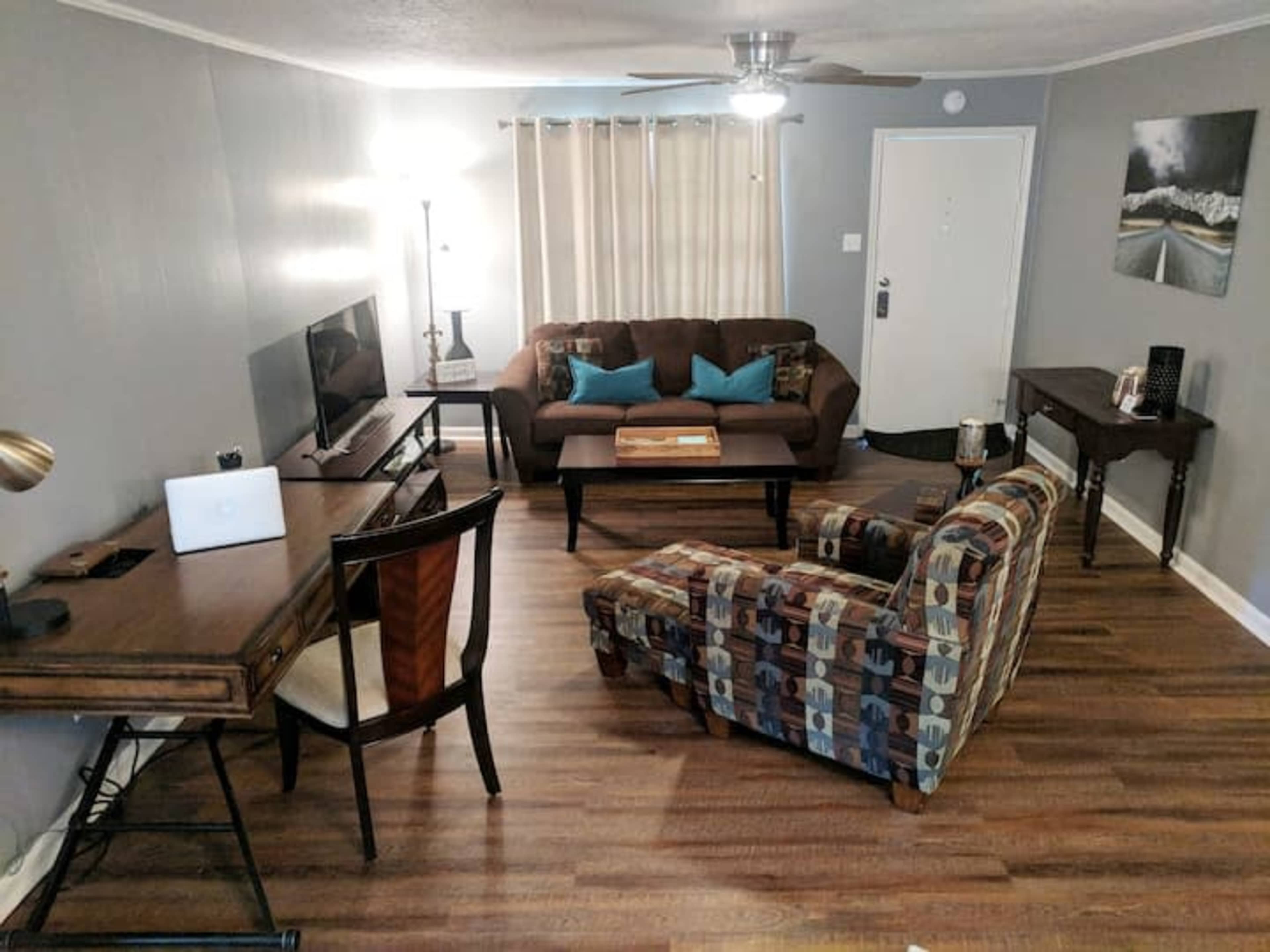 A living room featuring a brown couch, a chair with a patterned fabric, a wooden desk, and a television on a stand, all positioned around a central coffee table.