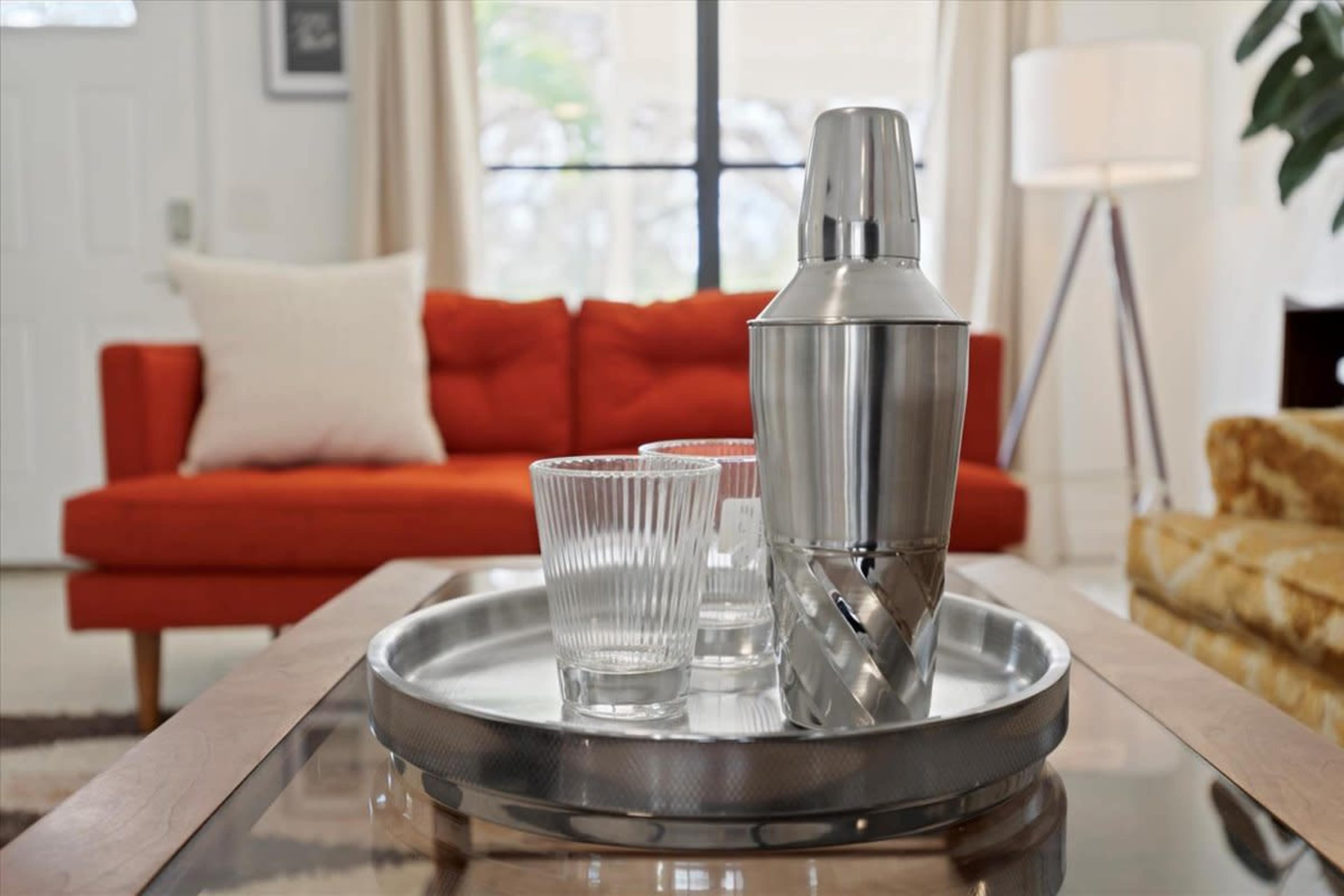 The image shows a living room with an orange couch, a silver cocktail shaker, and two clear glasses on a tray placed on a wooden coffee table.