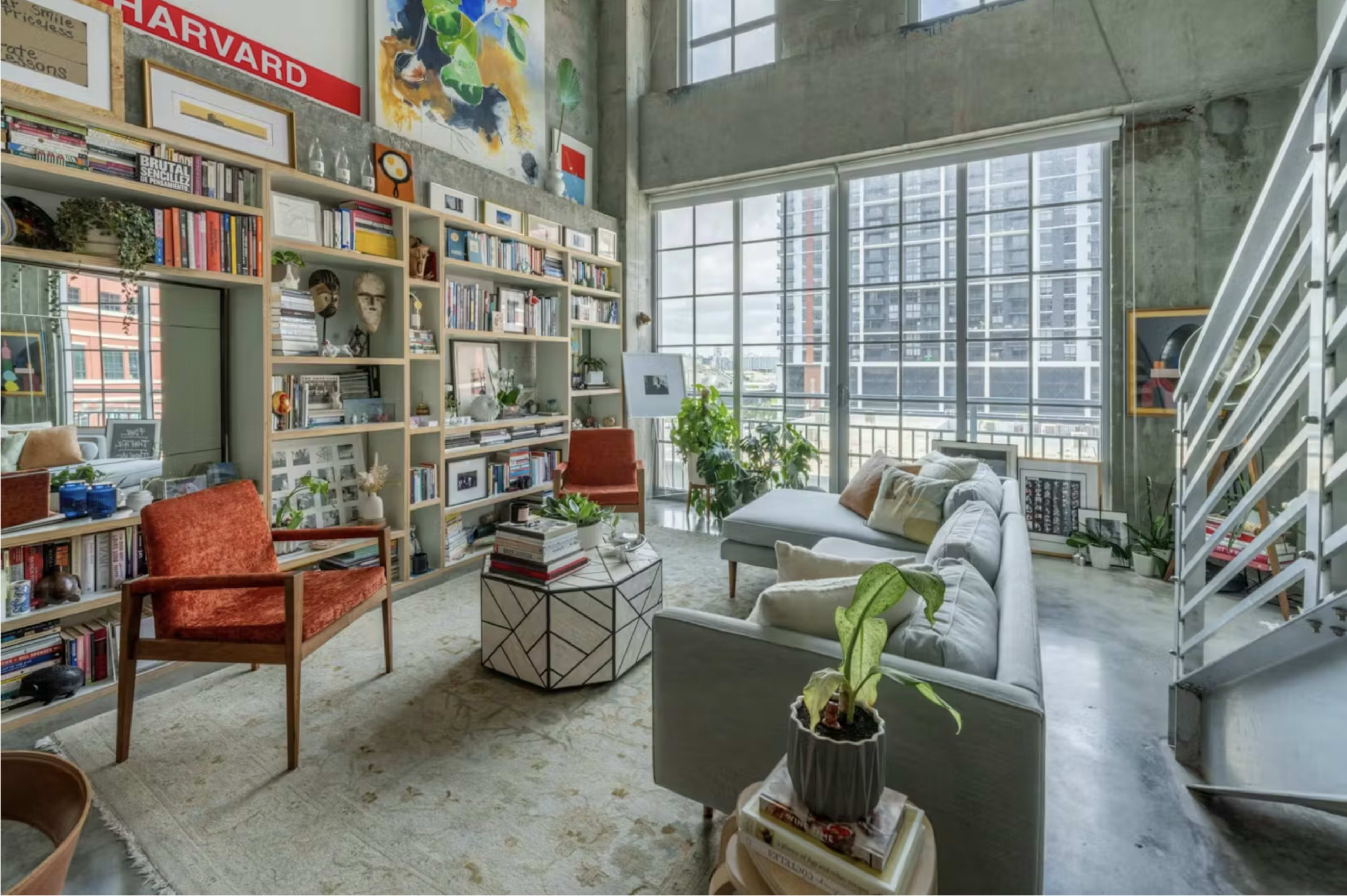A modern living room with large windows, bookshelves filled with books and framed pictures, and a mix of comfortable seating arrangements.