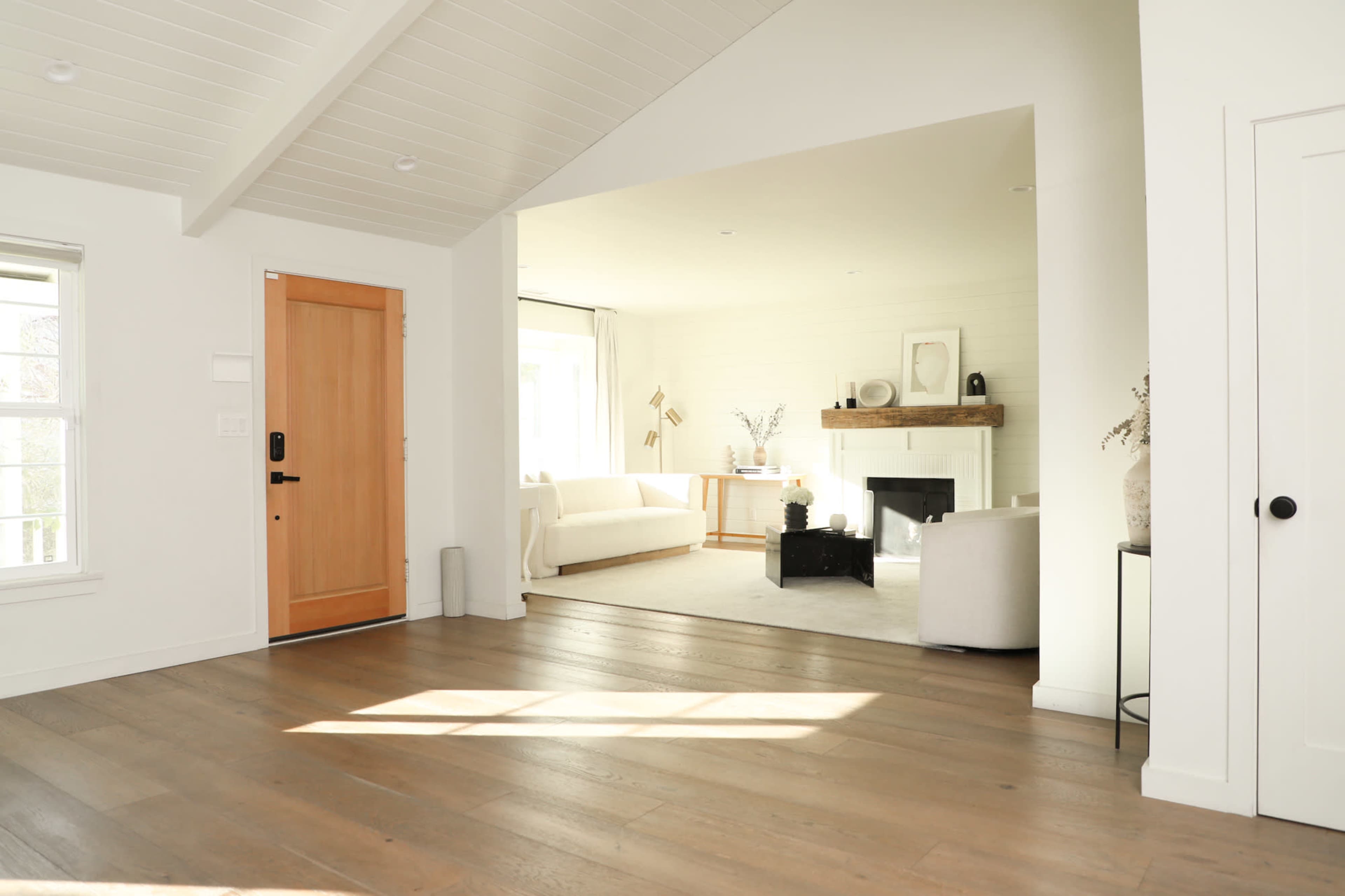 The image shows a bright, open interior space featuring a wooden front door, hardwood floors, and a living area with a white sofa and a fireplace.