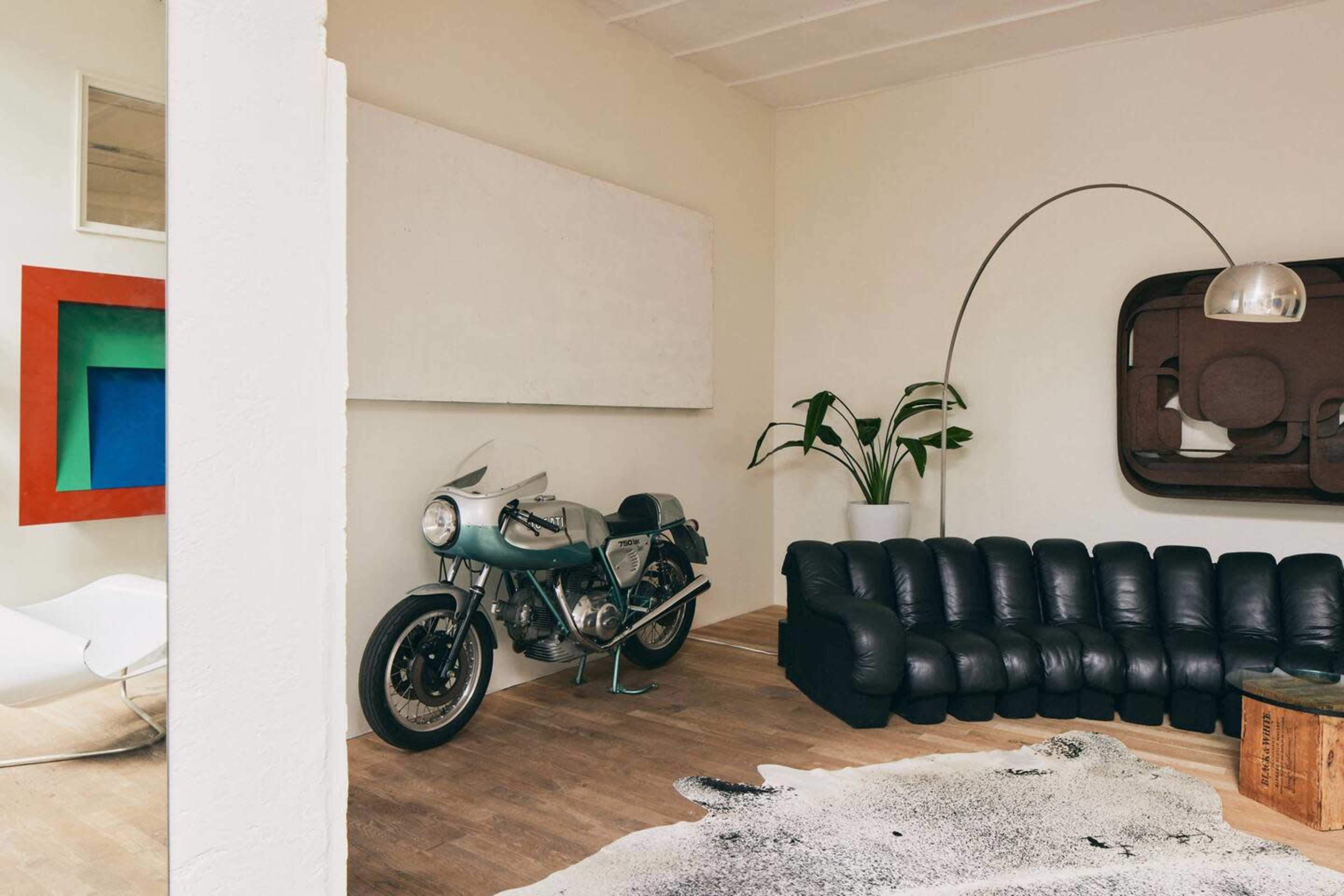 A modern living space with a black leather sofa, a motorcycle positioned against the wall, an oversized floor lamp, and abstract artwork mounted on the walls.