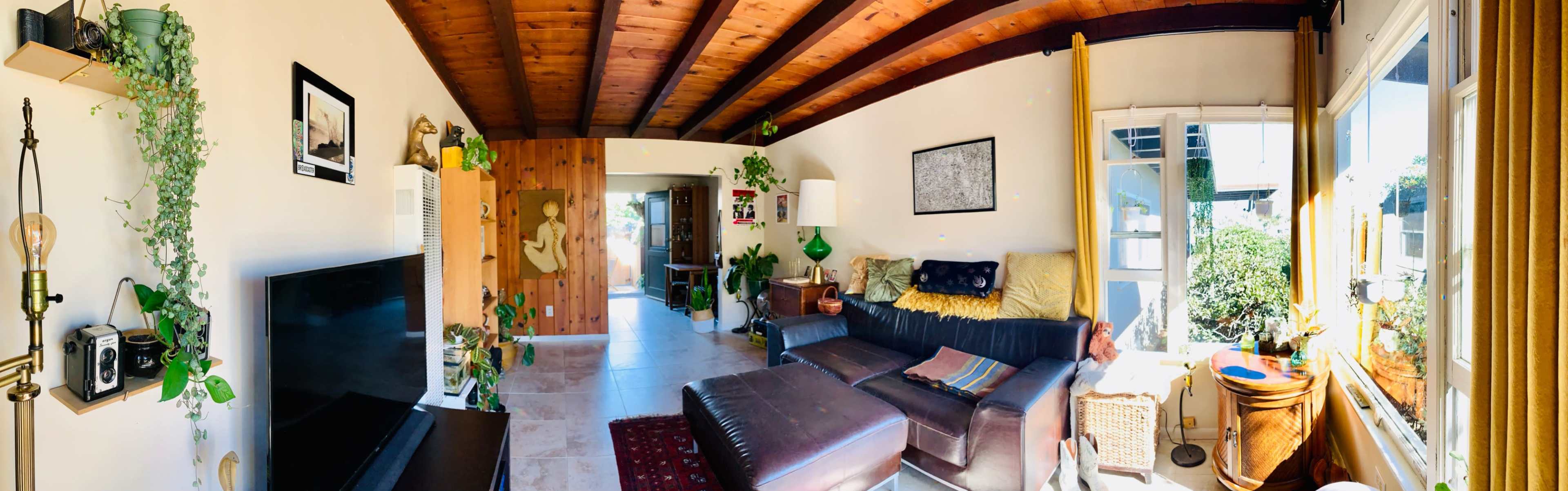 A spacious living room with a sectional couch, plants, artwork on the walls, and large windows allowing natural light to enter.