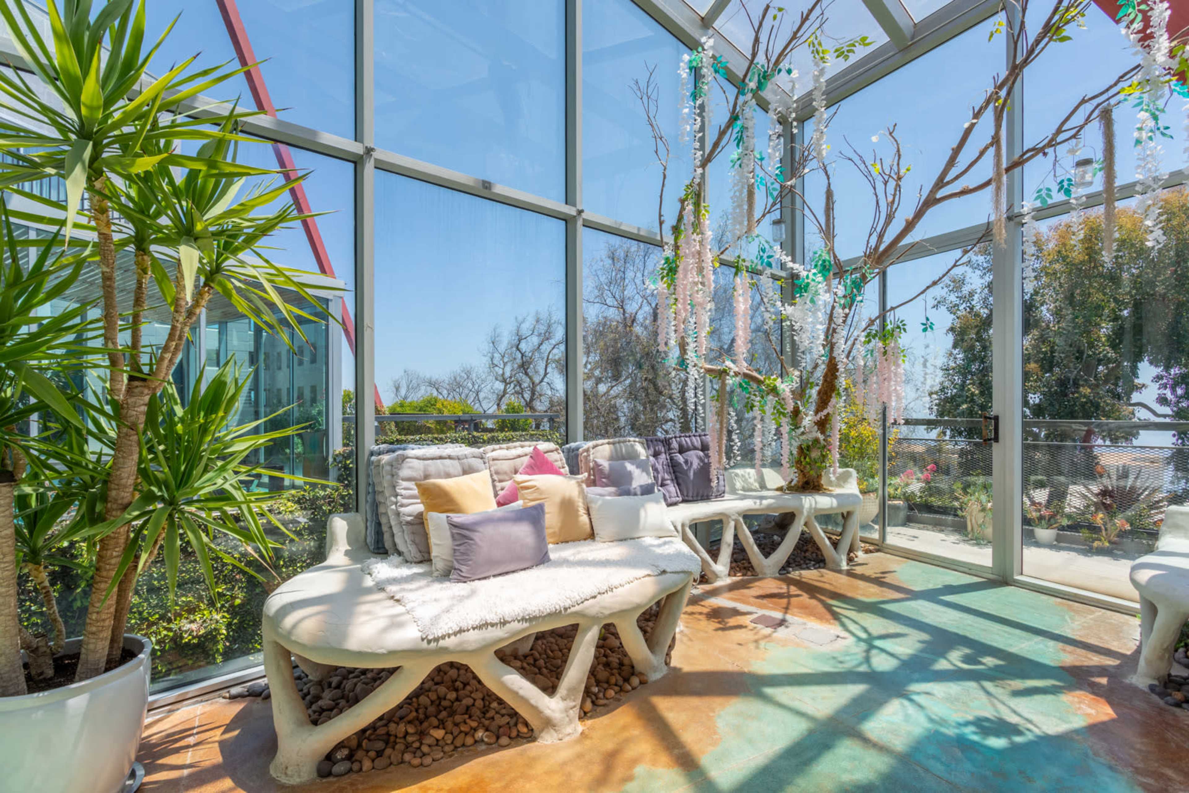 A bright sunroom features white modern seating arrangements surrounded by greenery and playful decor, with large glass walls allowing natural light to flood the space.