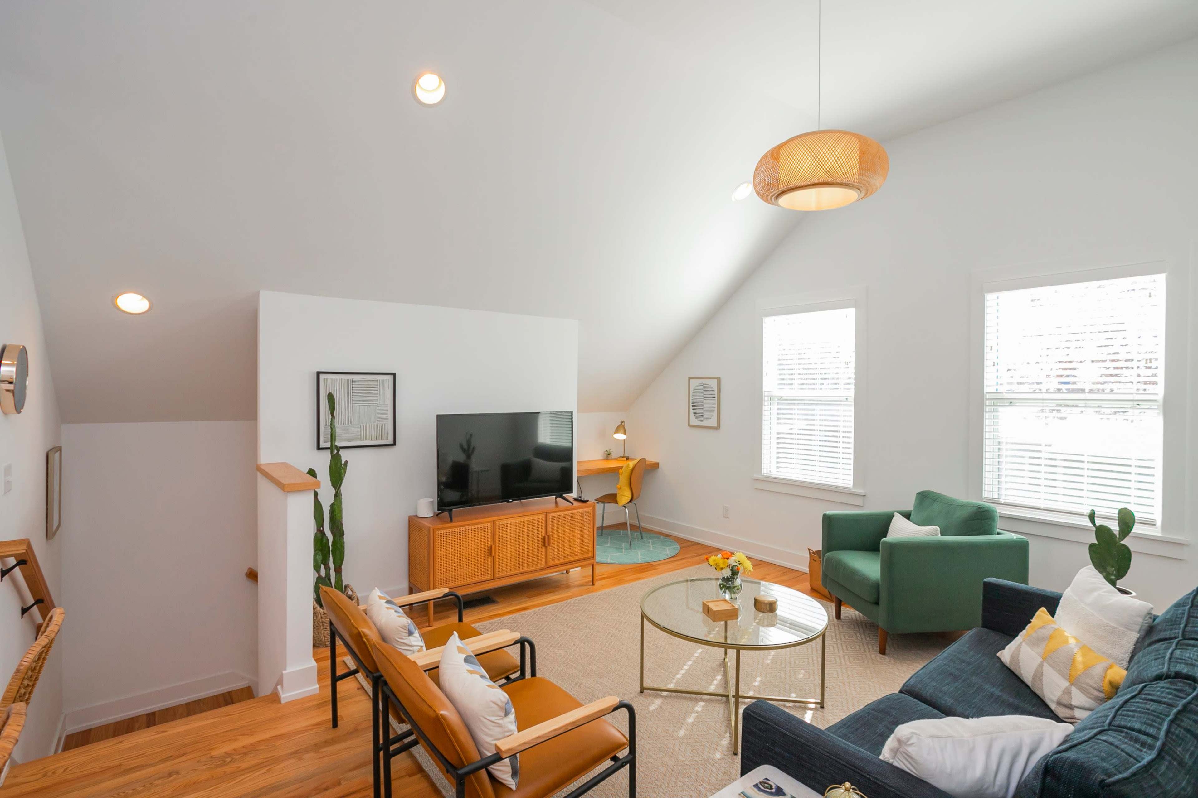 The image shows a bright, modern living room with a television, a coffee table, and various seating arrangements, illuminated by natural light through multiple windows.