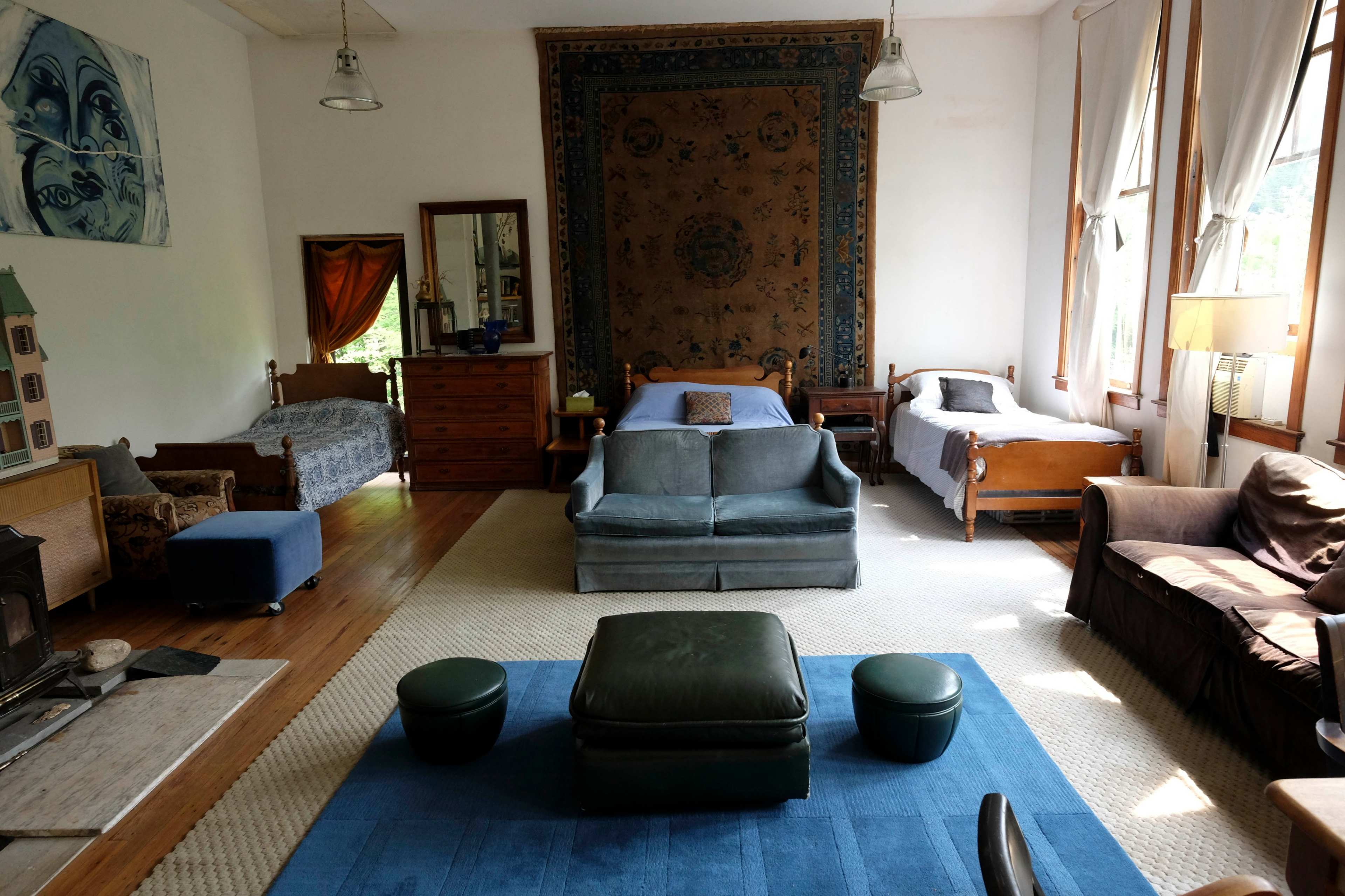 The room features a mix of seating options, two beds, a decorative wall tapestry, and large windows allowing natural light.