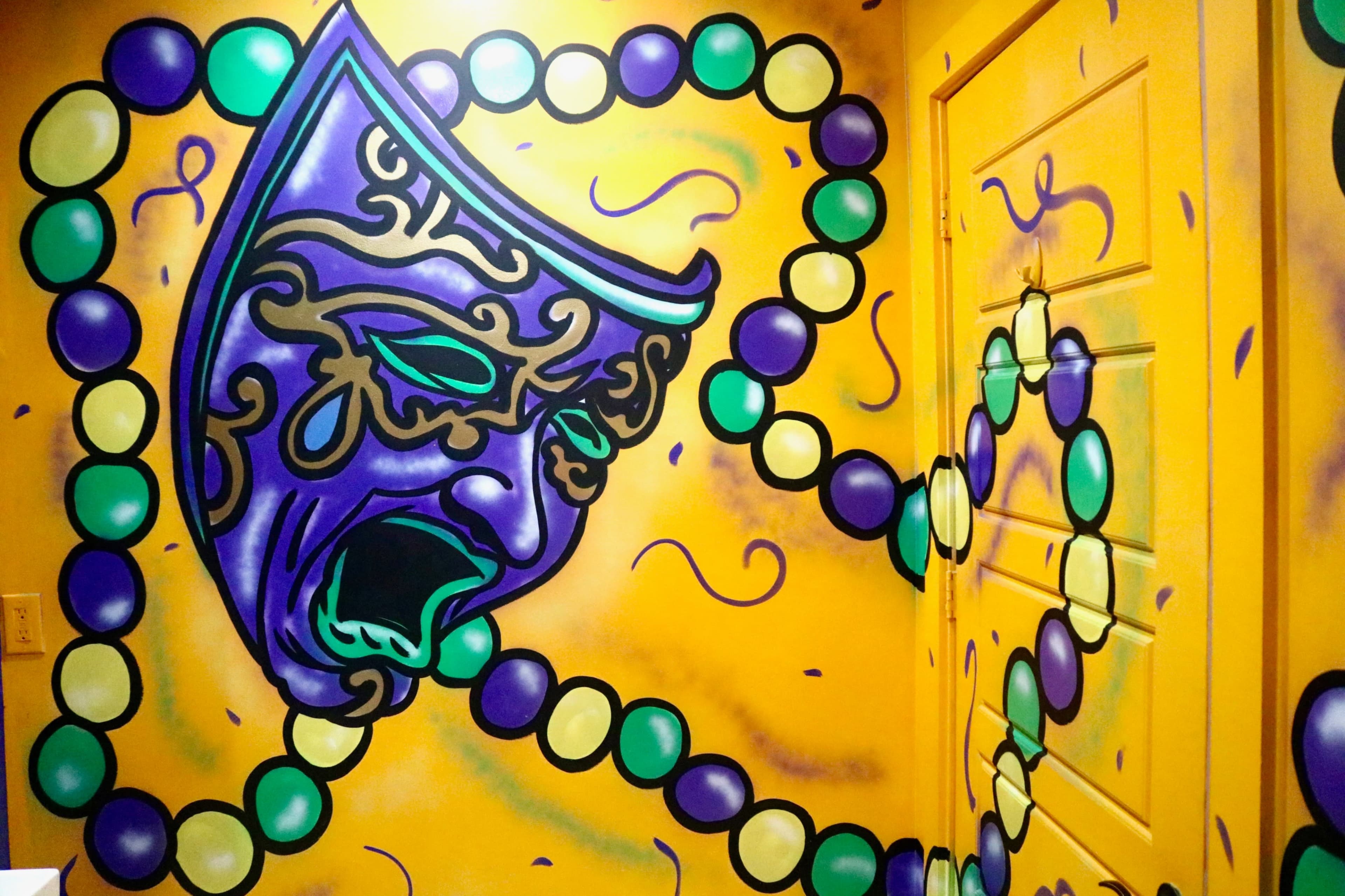 A vibrant mural featuring a dramatic mask and colored beads is painted on a yellow wall next to a door.