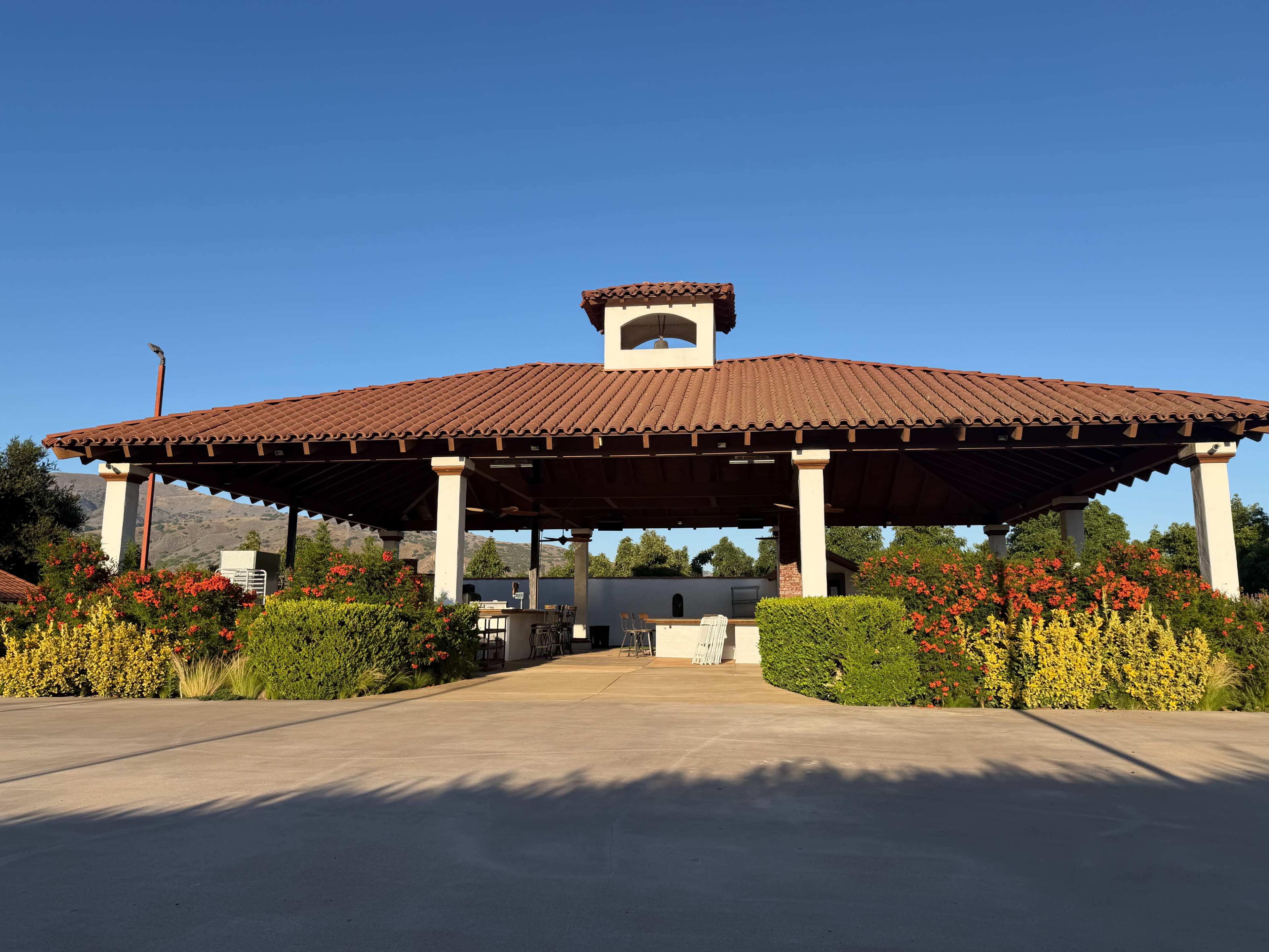 A large, open pavilion with a tiled roof stands surrounded by colorful shrubs and flowers against a clear blue sky.