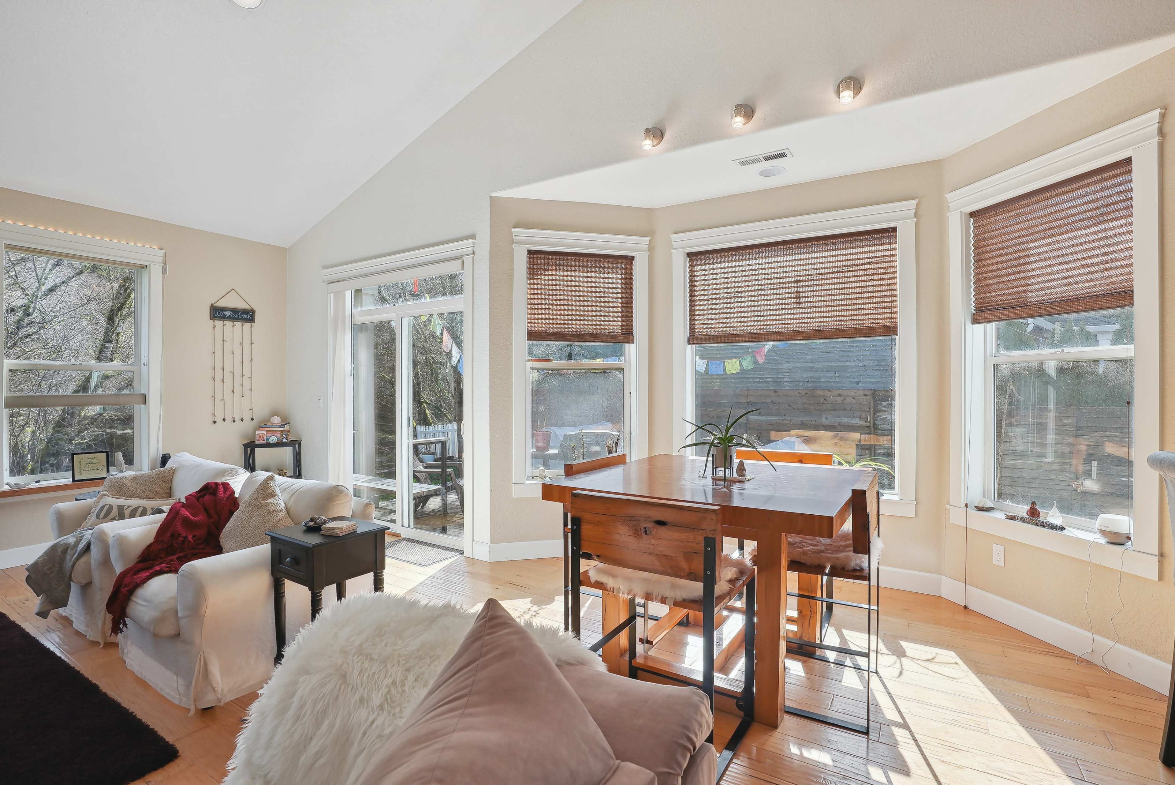 A sunlit living space features a dining table near large windows adorned with wooden blinds, surrounded by cozy furniture and a view of the outdoors.