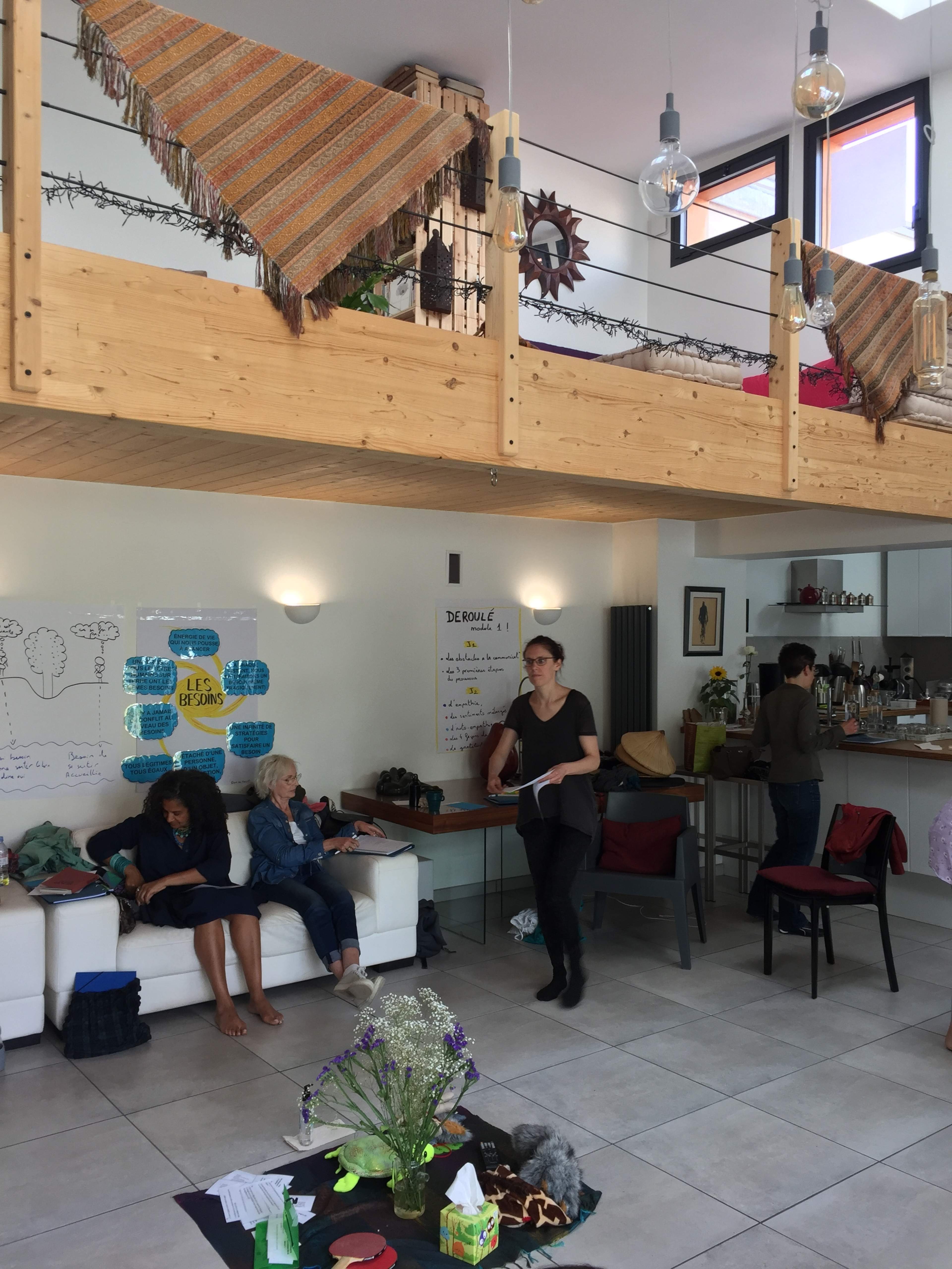 An open living space with a lofted area, where people are seated and engaged in various activities, surrounded by decorative elements and plants.