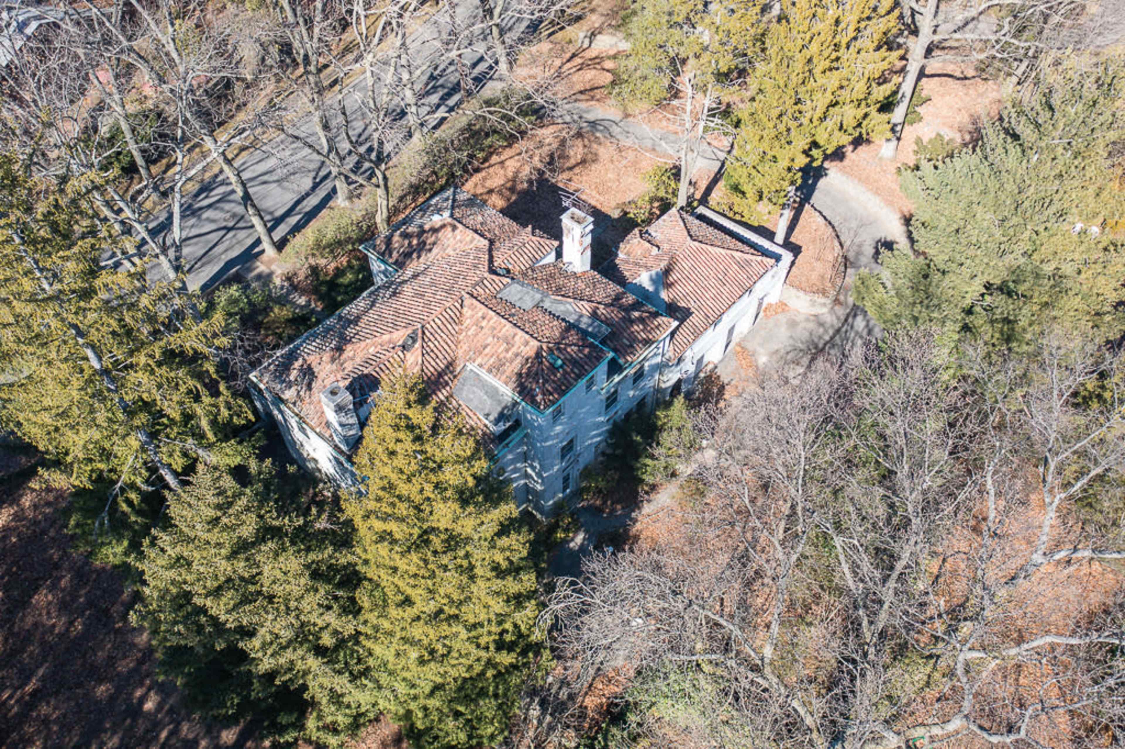 An aerial view shows a single-story house with a tiled roof surrounded by trees and a winding path in a residential area.