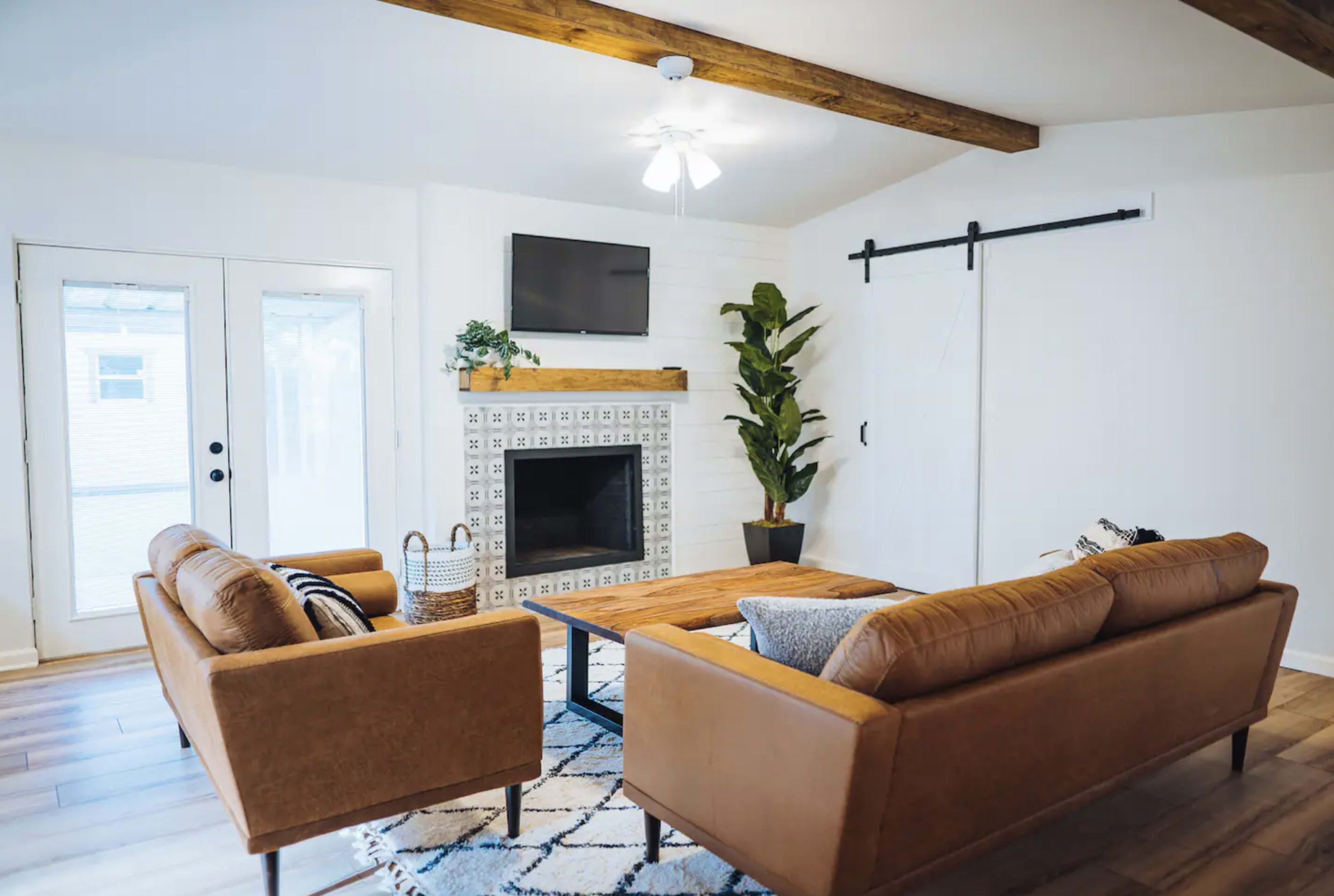 The living room features two brown sofas facing a wooden coffee table in front of a brick fireplace, with a large window and barn door visible on the left.