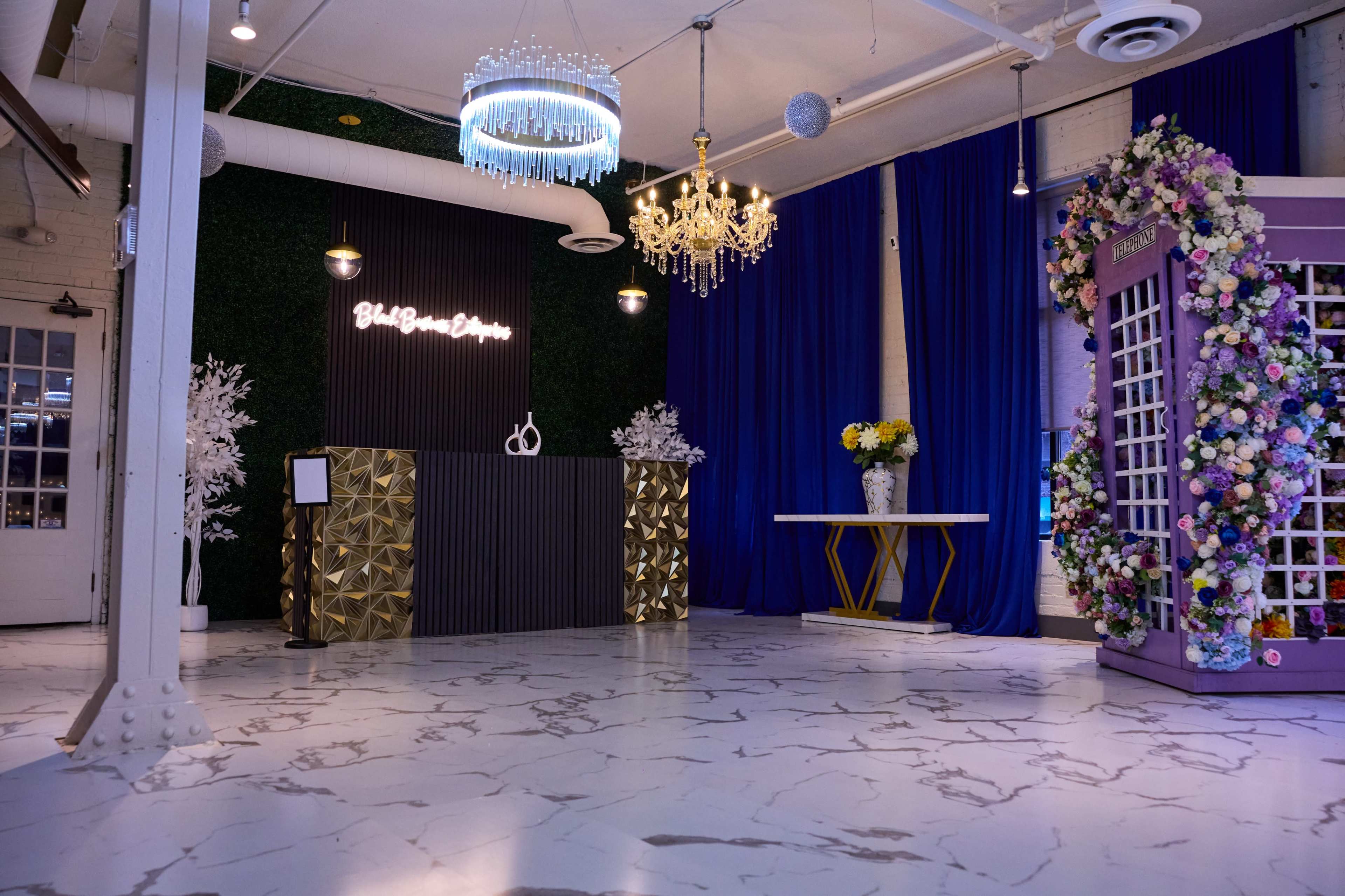 The image shows a decorated event space featuring a reception desk, elegant lighting, a floral arch, and a table set against lush greenery and blue drapes.