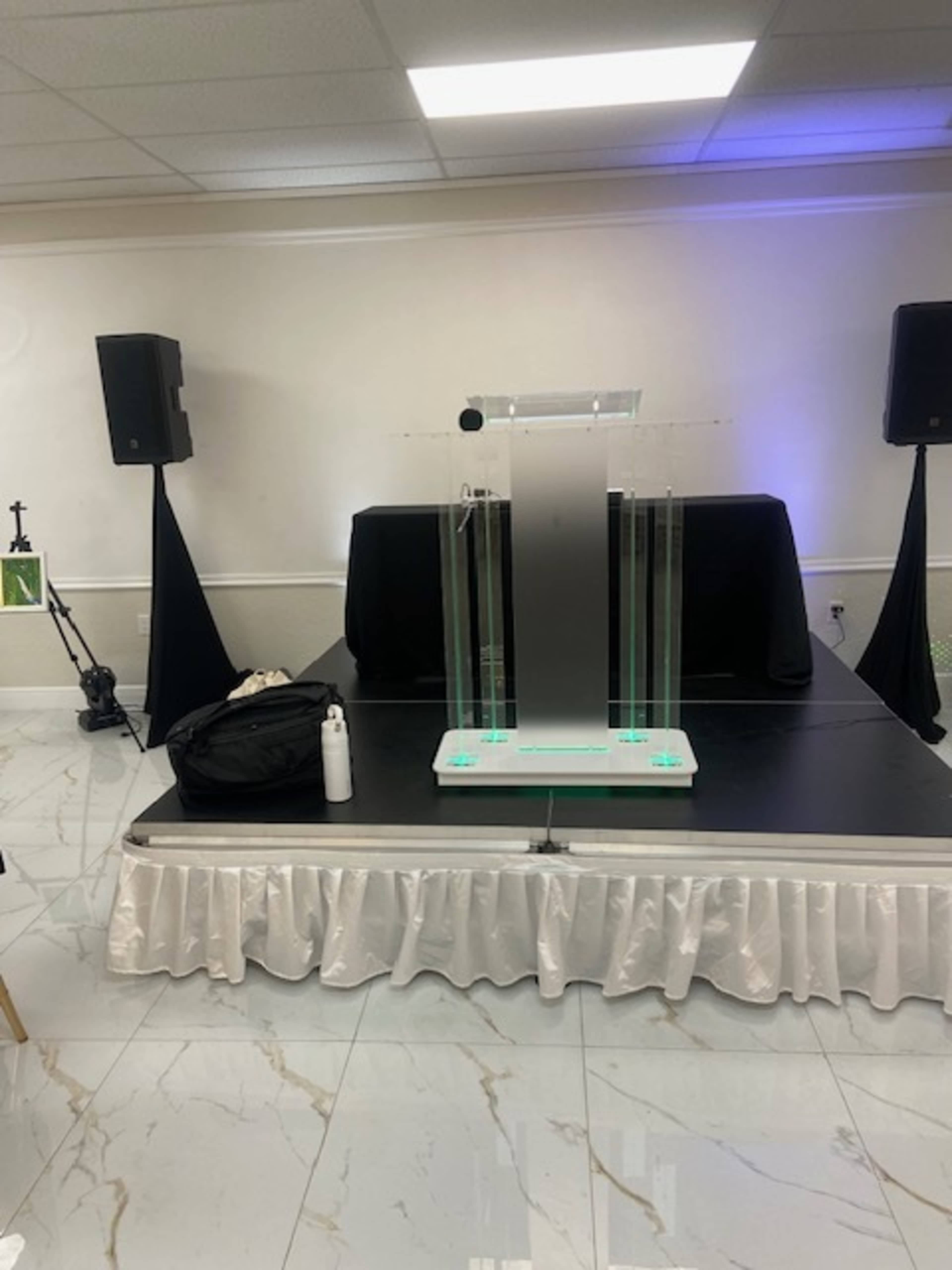 A podium with a transparent design is set on a stage in a room featuring speaker stands and a black backdrop.
