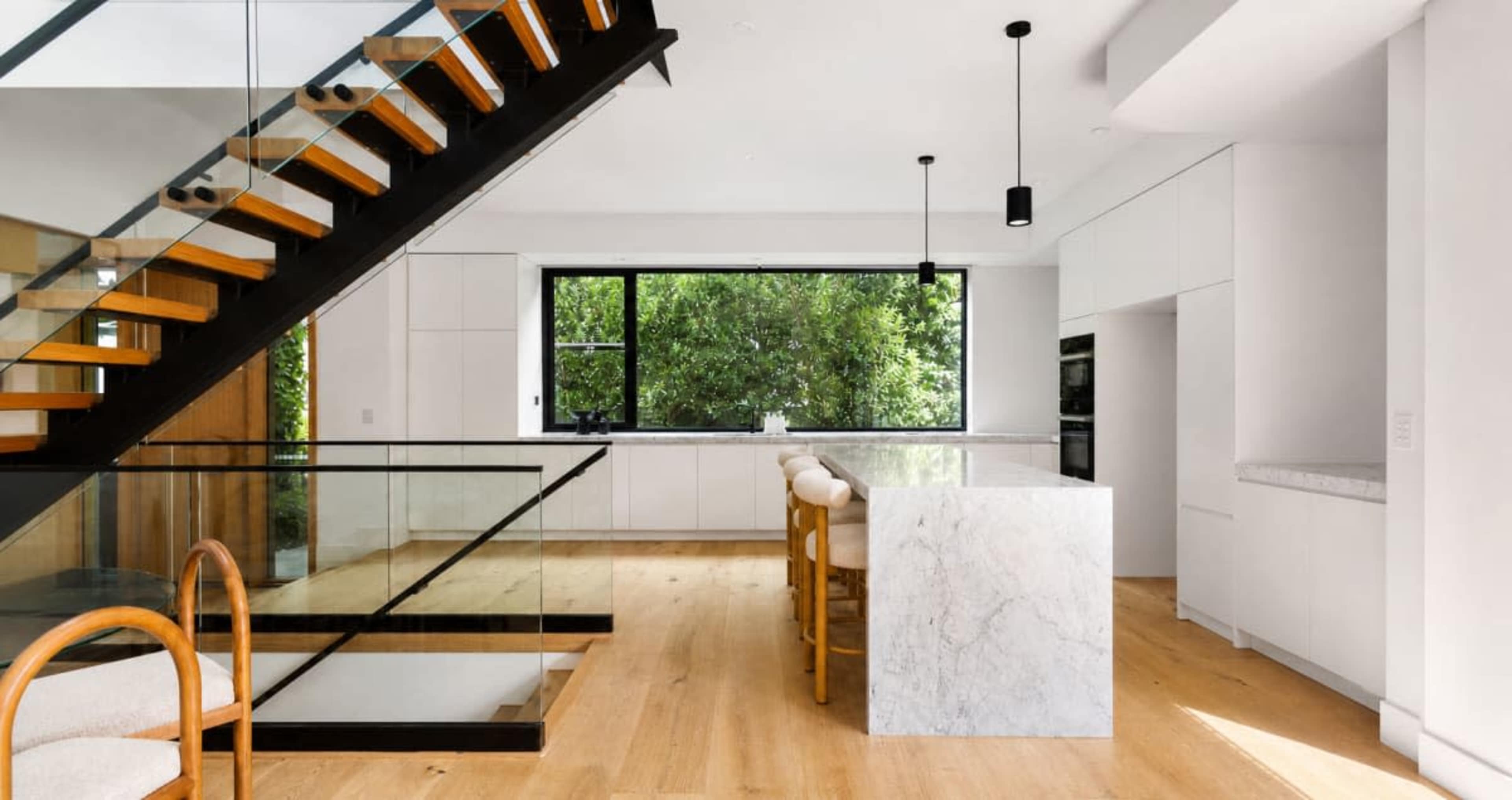 The image shows a modern kitchen with a marble island, minimalist cabinetry, and a staircase with wooden treads and glass railings.
