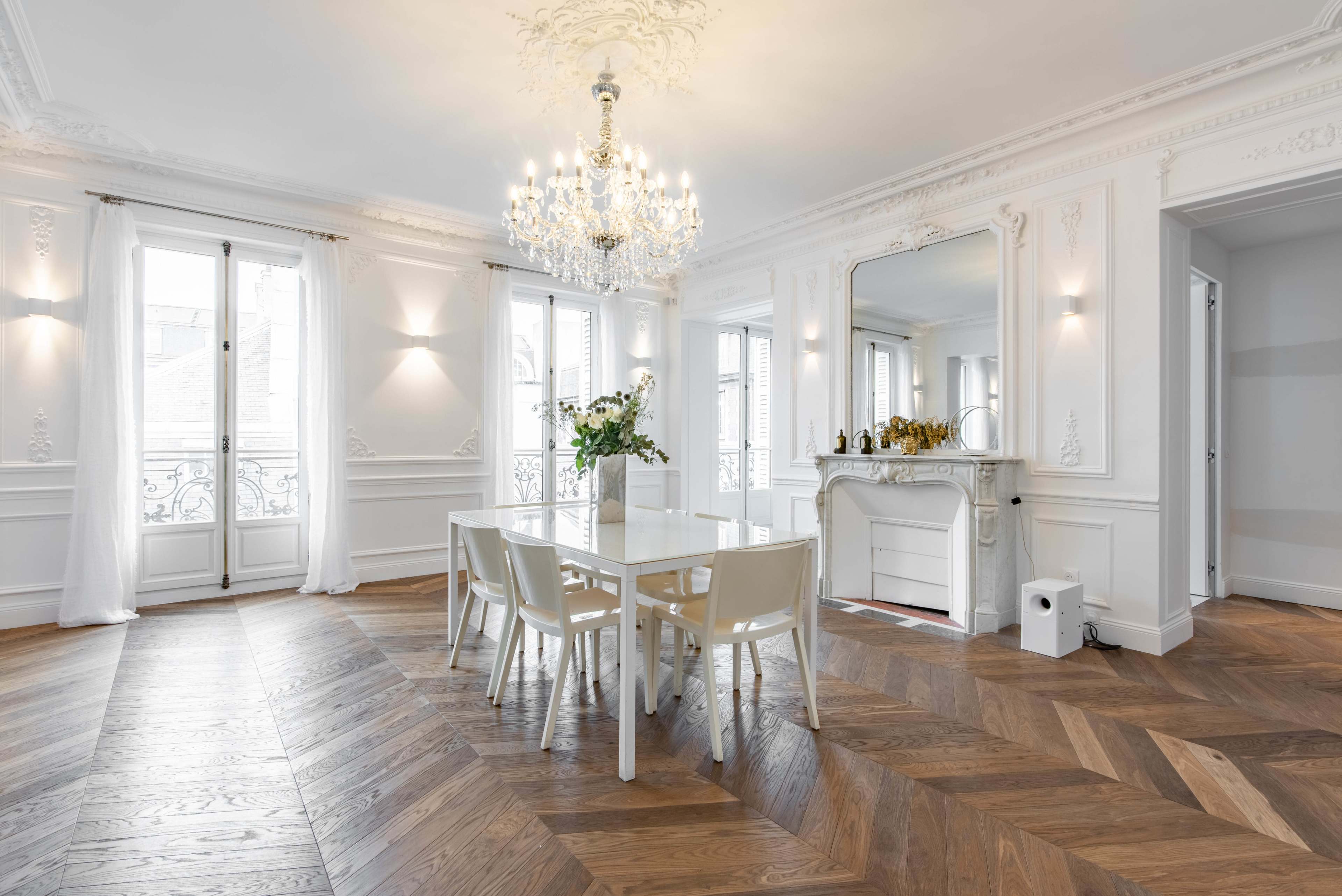 A bright dining area features a glass table surrounded by white chairs, with a chandelier overhead and ornate details on the walls.