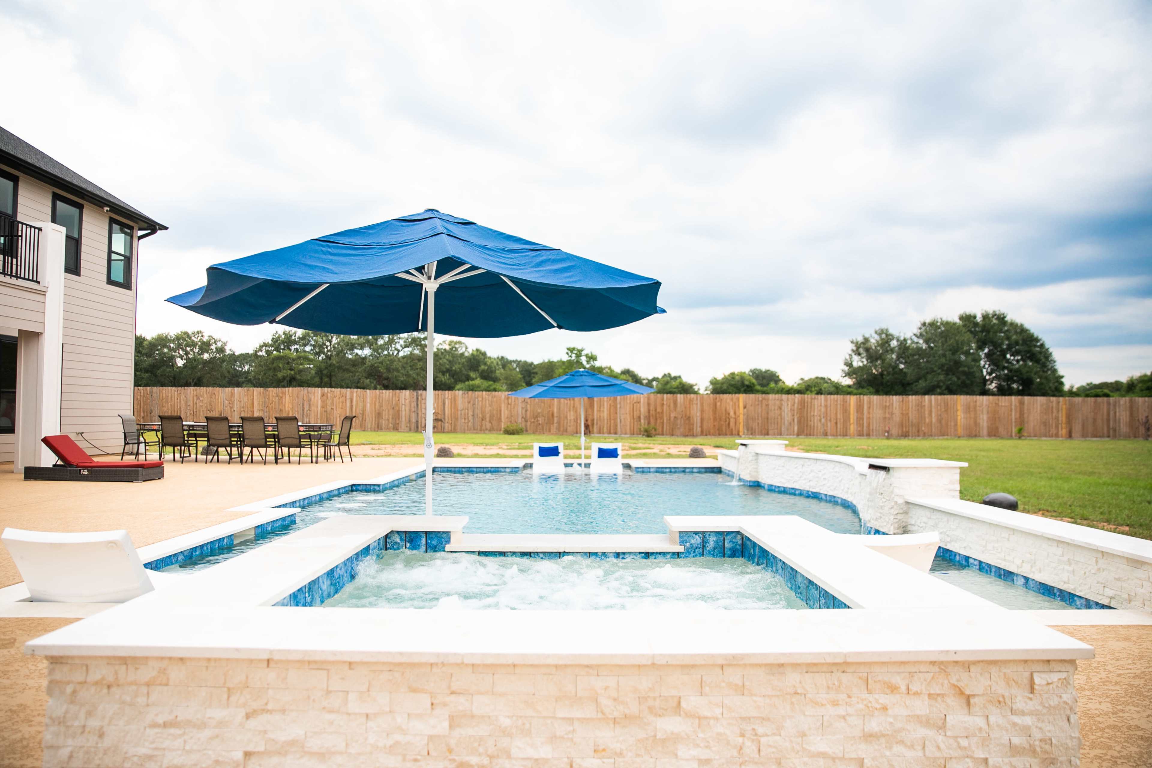 A swimming pool with a spa is surrounded by a patio, featuring a large blue umbrella and a dining set in the background.