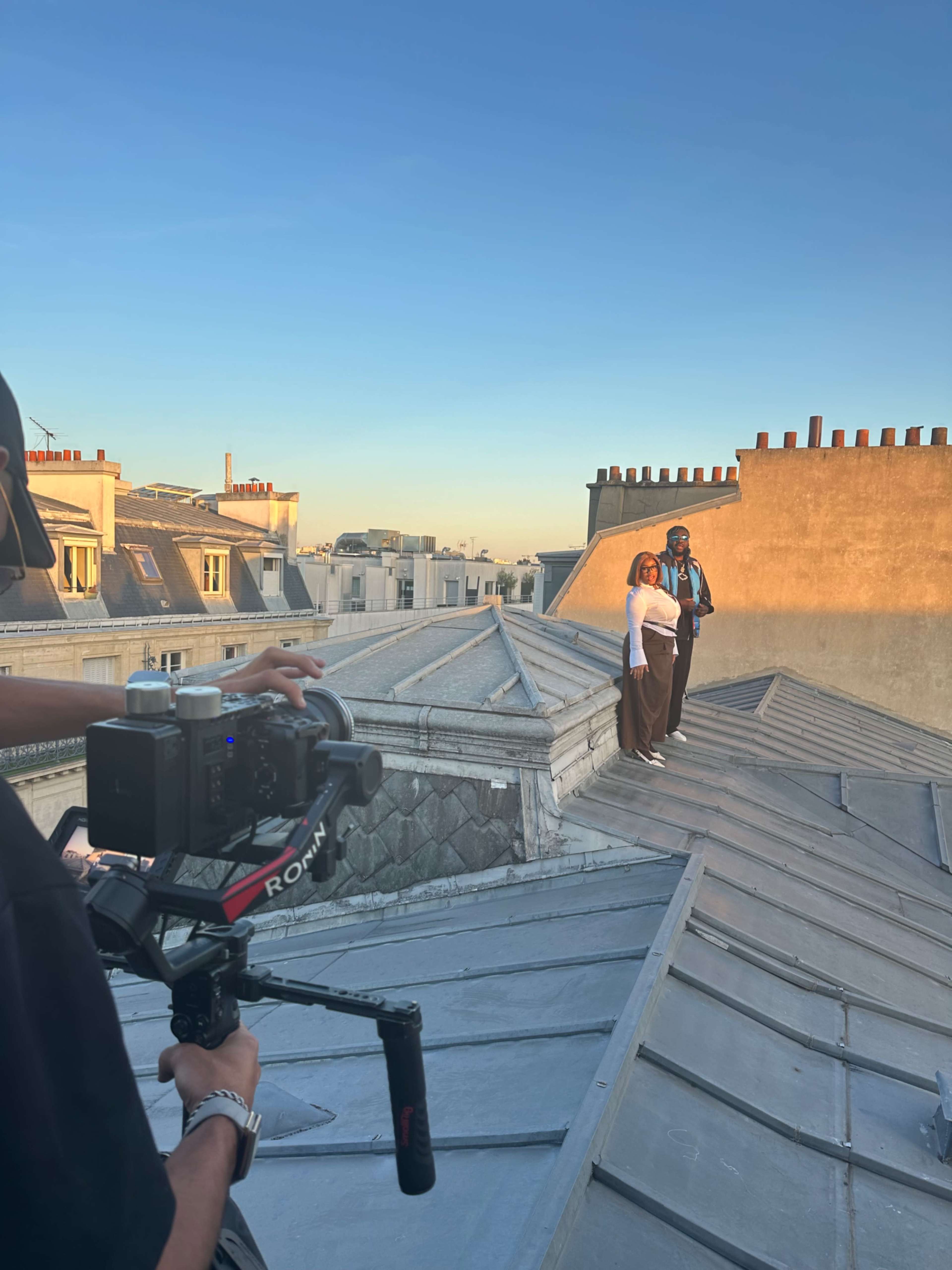 A photographer captures a photo shoot on a rooftop with two models against a clear blue sky and city skyline.