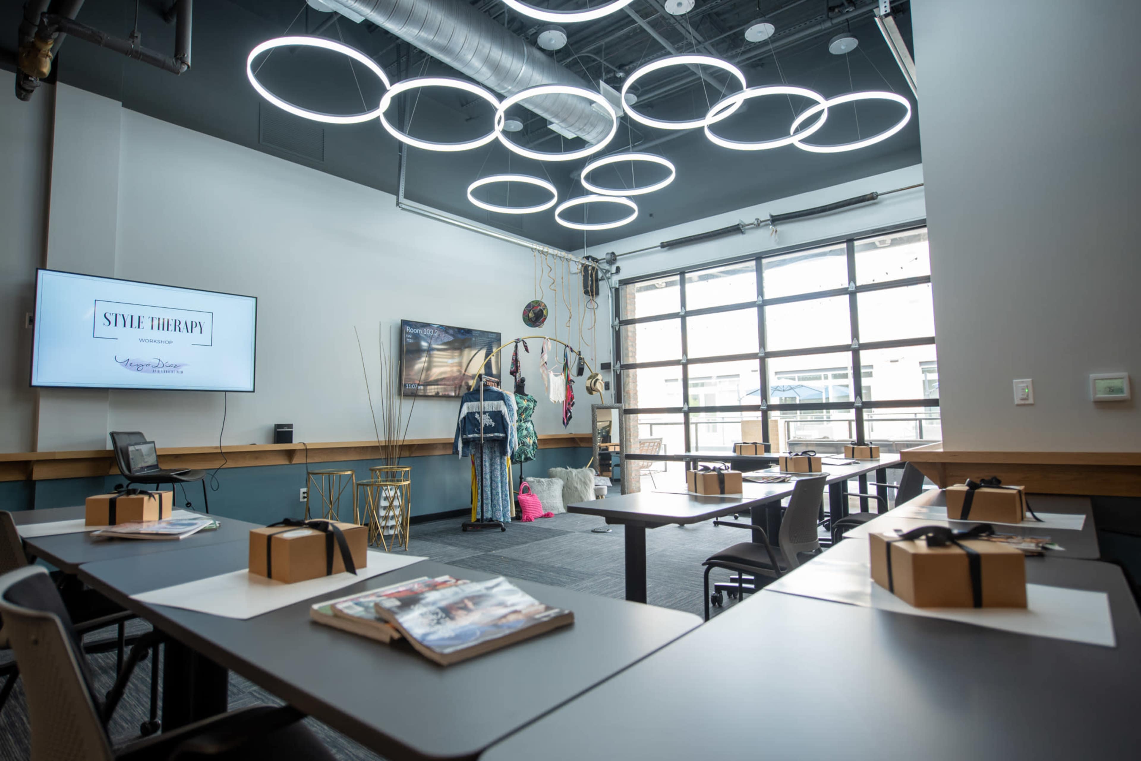 The image shows a modern interior space designed for a style therapy session, featuring a presentation screen, a TV, multiple tables set up with materials, and unique circular light fixtures on the ceiling.