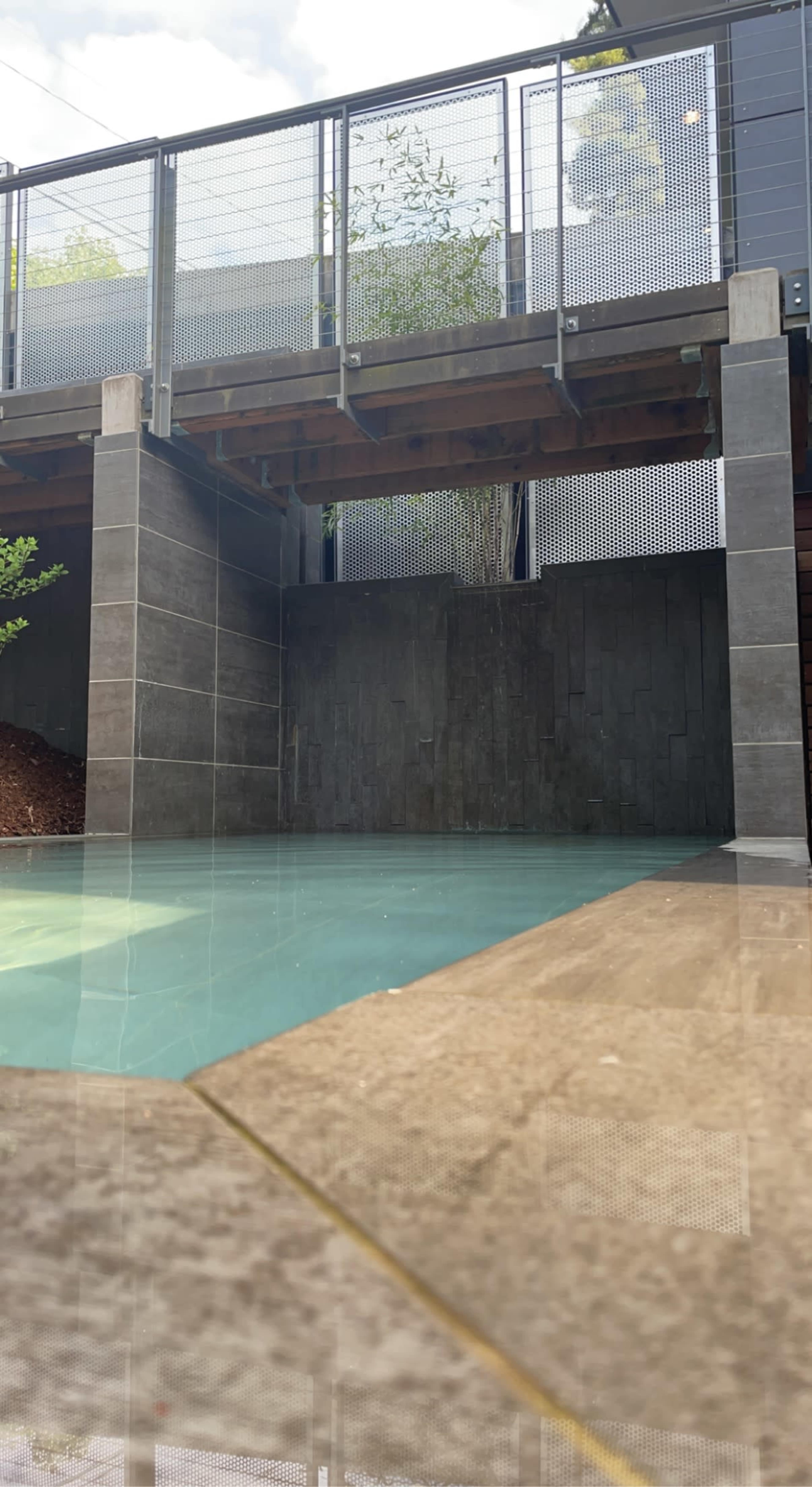 A modern pool area with a concrete structure overhead, surrounded by stone walls and a wooden deck.