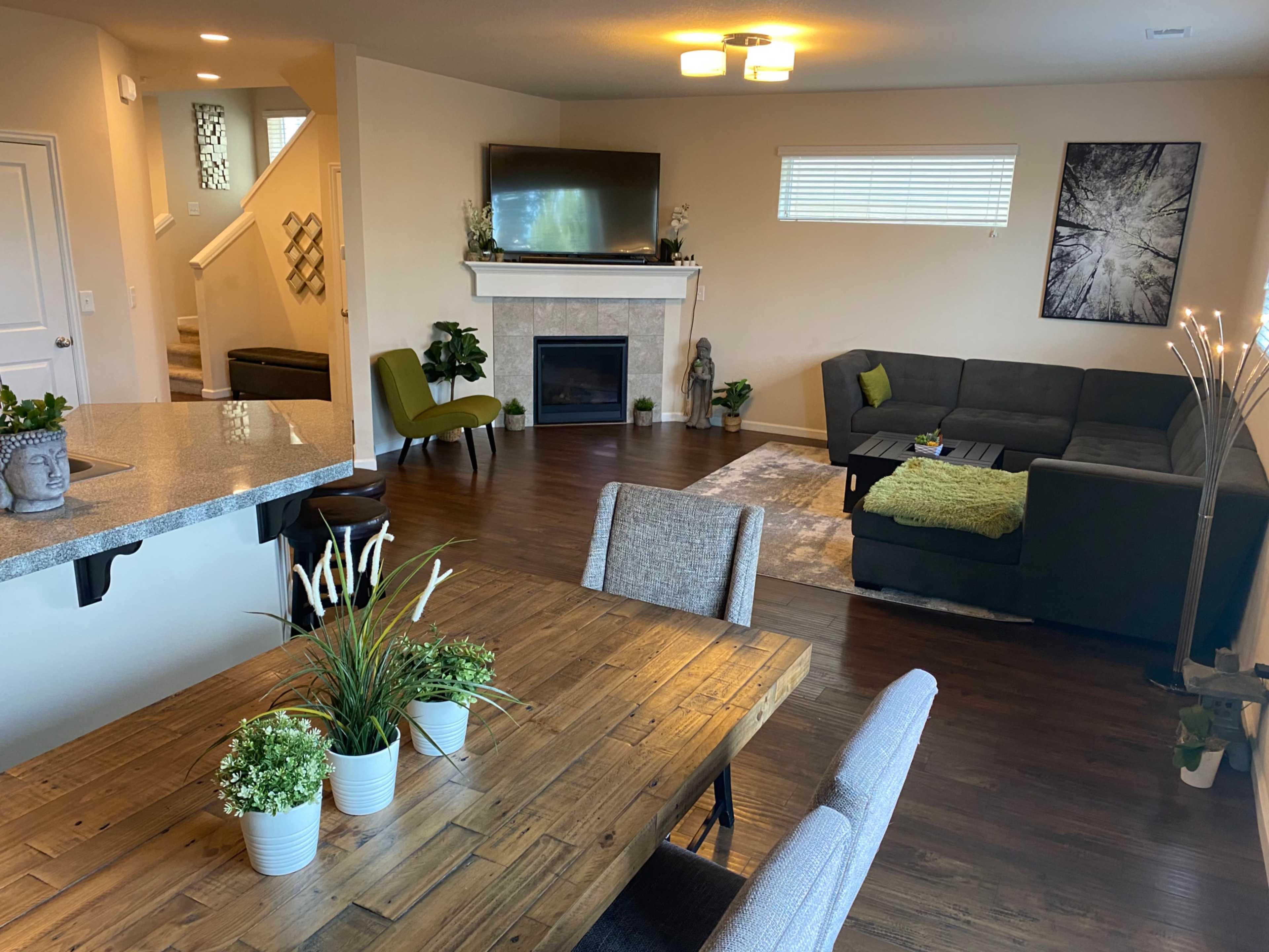 A modern living room and dining area featuring a gray sectional sofa, a fireplace, a dining table with plants, and a television mounted on the wall.