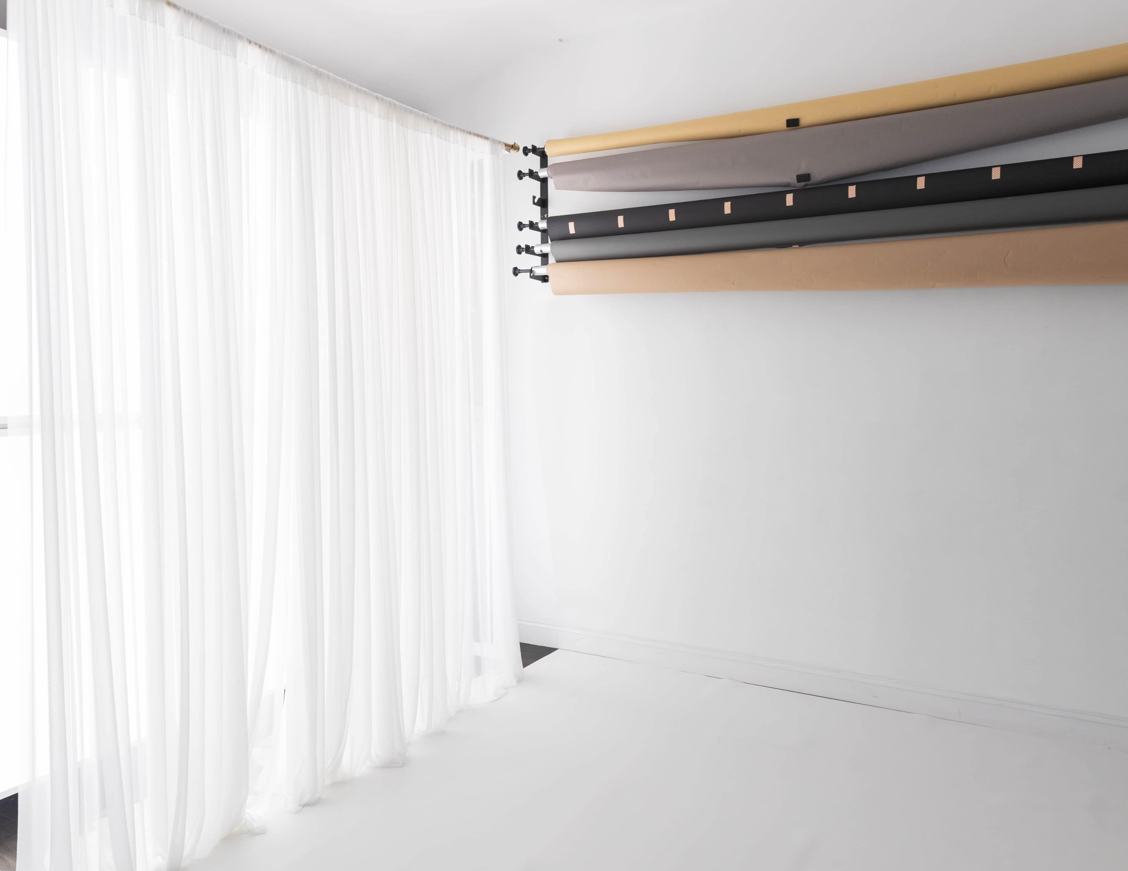 The image shows a bright, minimalistic studio space with sheer white curtains and a backdrop stand holding various rolls of colored backdrop paper.