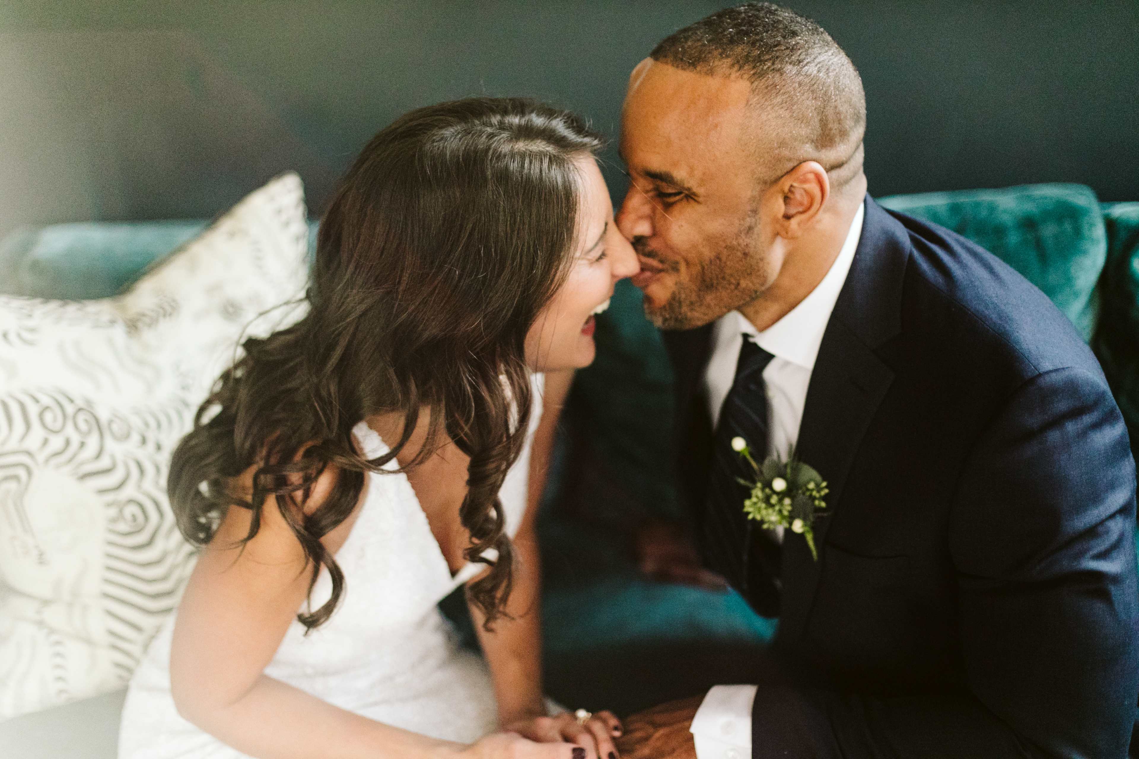 A bride and groom share a joyful moment close together on a couch, both smiling as they touch noses.