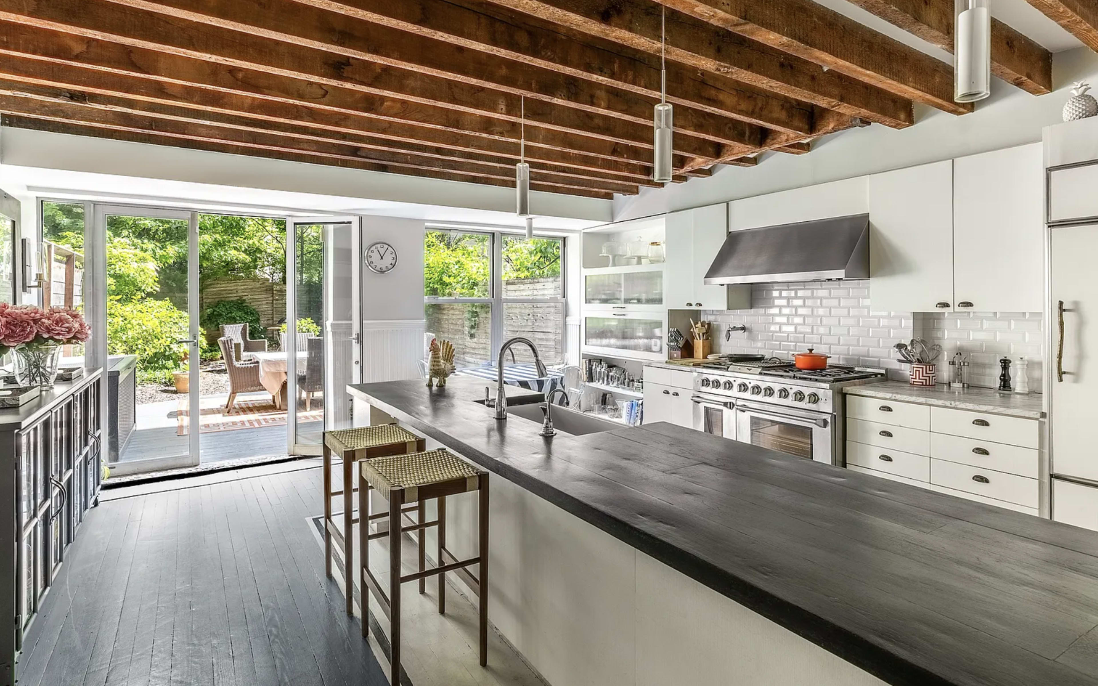 The image shows a modern kitchen with a large island, stainless steel appliances, and exposed wooden beams, leading to a patio area visible through a sliding glass door.