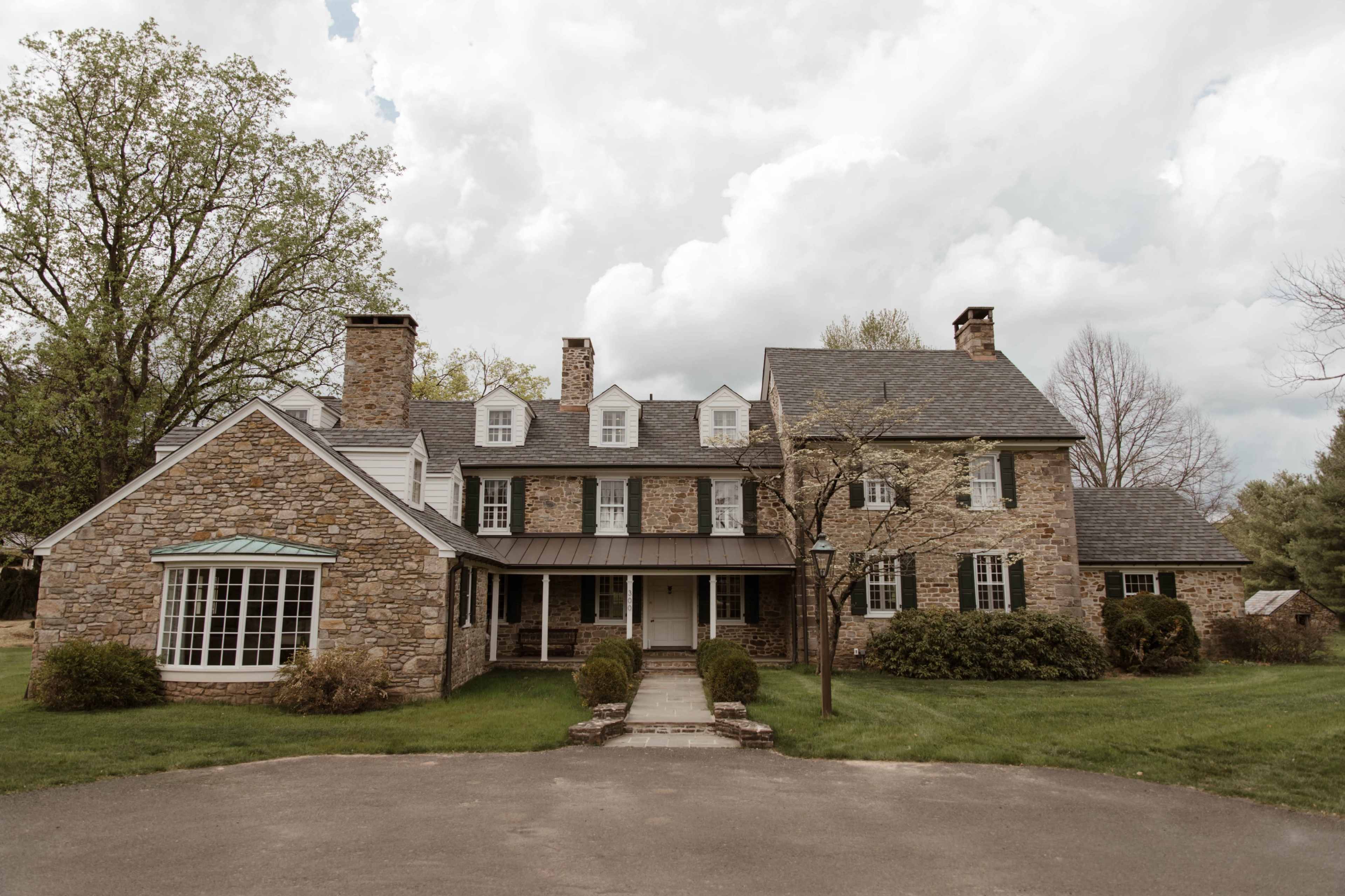A large stone house with multiple gabled roofs, a front walkway, and surrounding greenery under a cloudy sky.